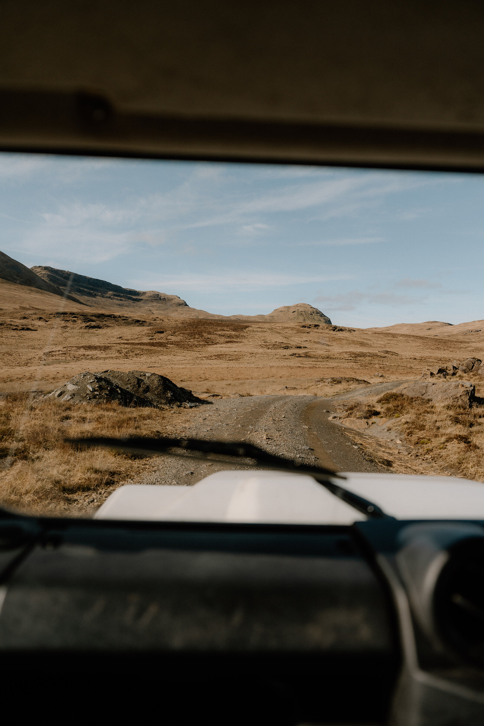 A dirt track winds through rocky moorland beneath distant mountains on the Isle of Rum, seen through a vehicle's windscreen.