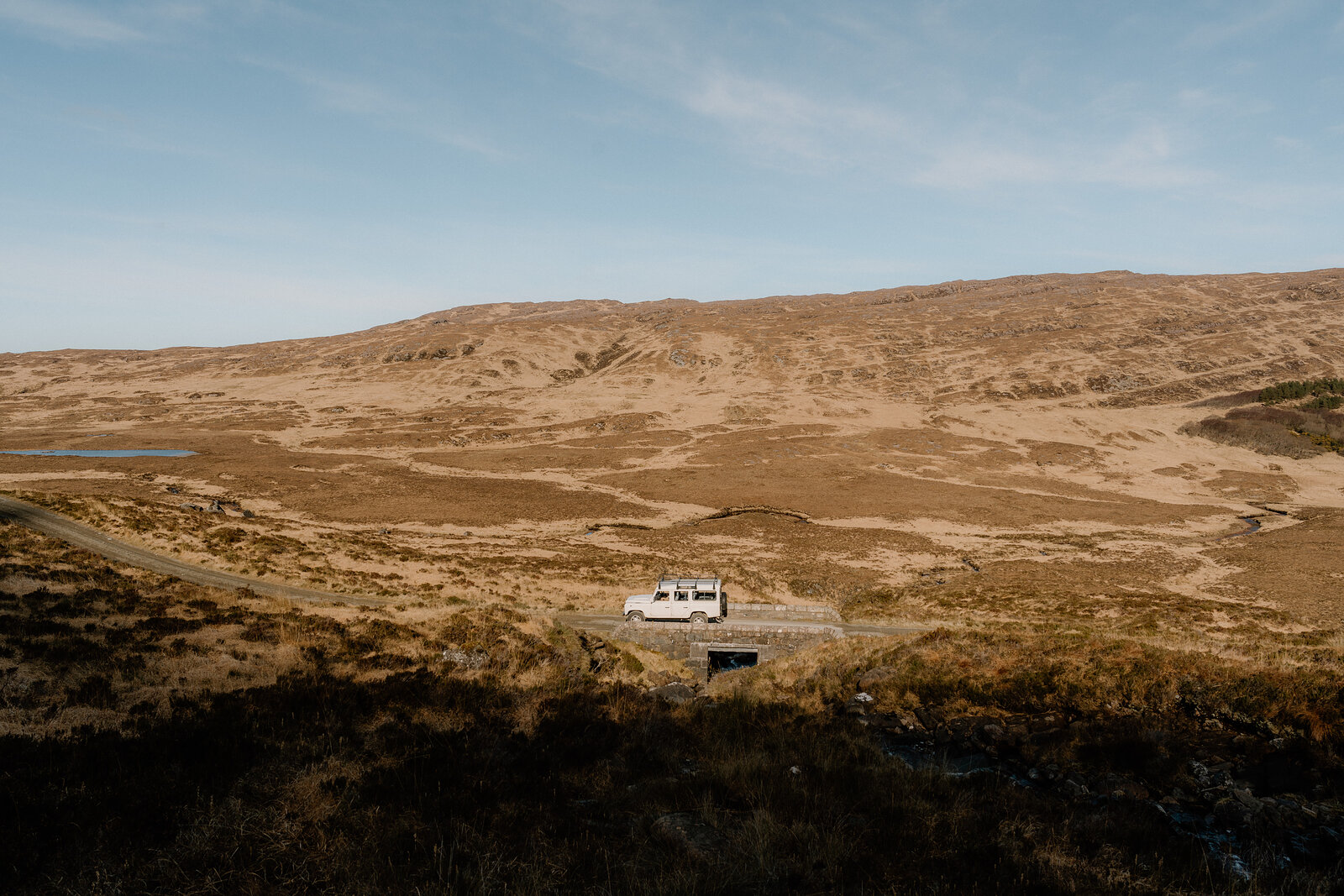 A white Land Rover is parked on a dirt track crossing open moorland on the Isle of Rum, a small loch visible in the distance.