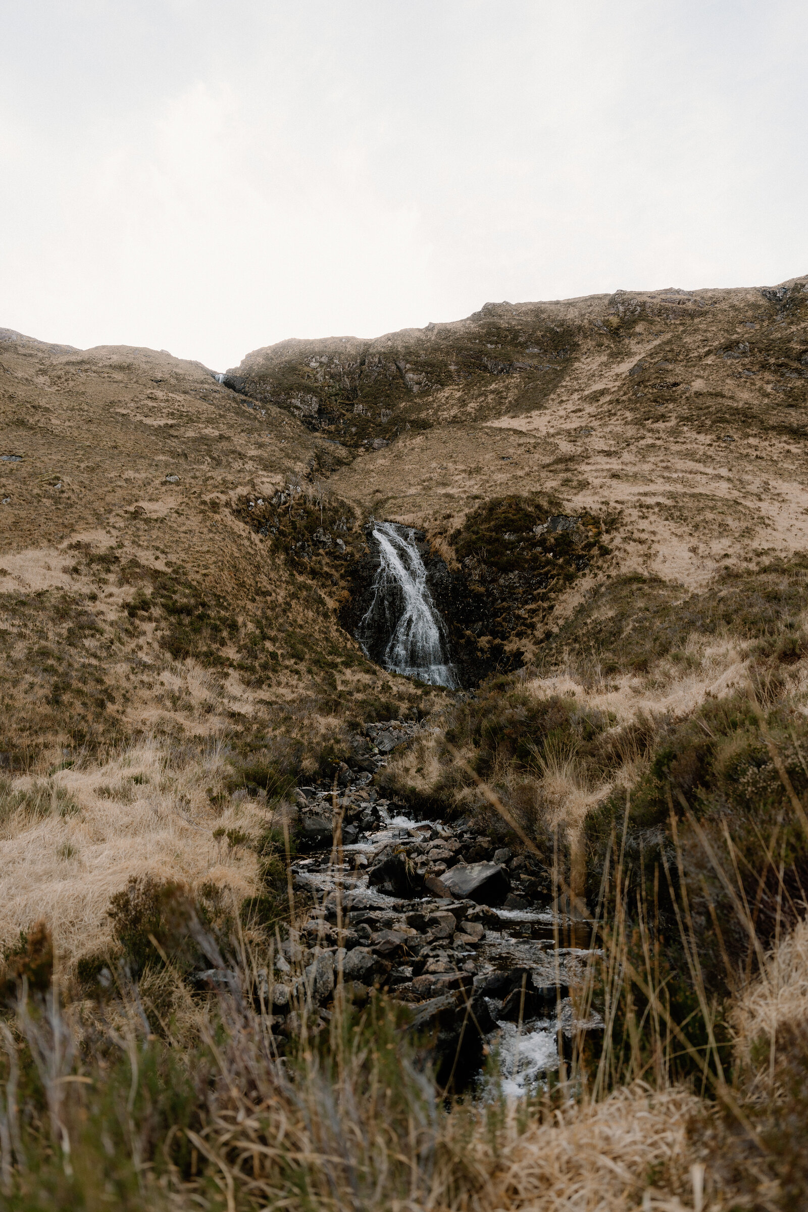 A waterfall tumbles down a rocky gully on the Isle of Rum, a stream winding through grass below.