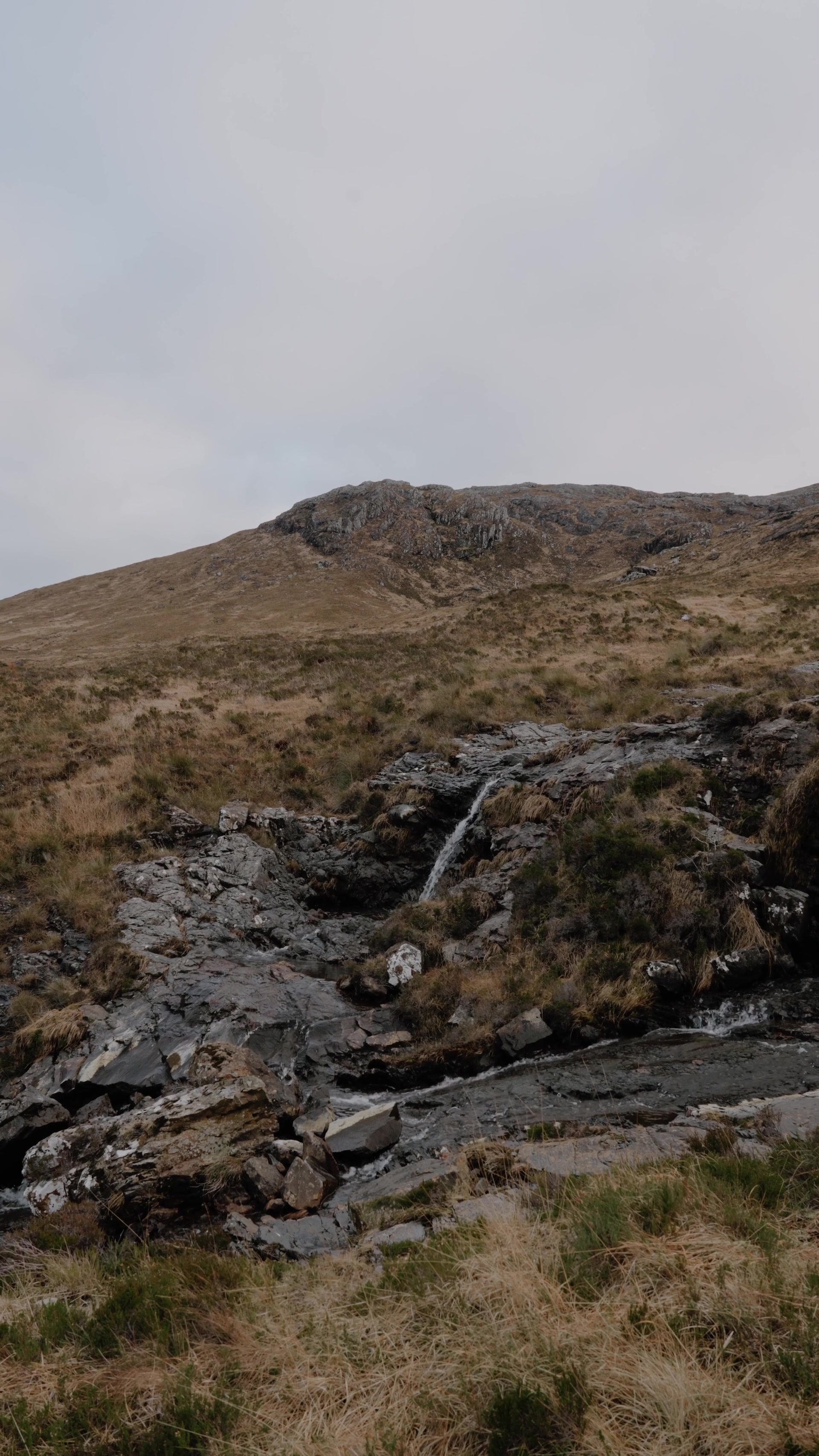A small burn cascades down a rocky hillside below a rugged peak.