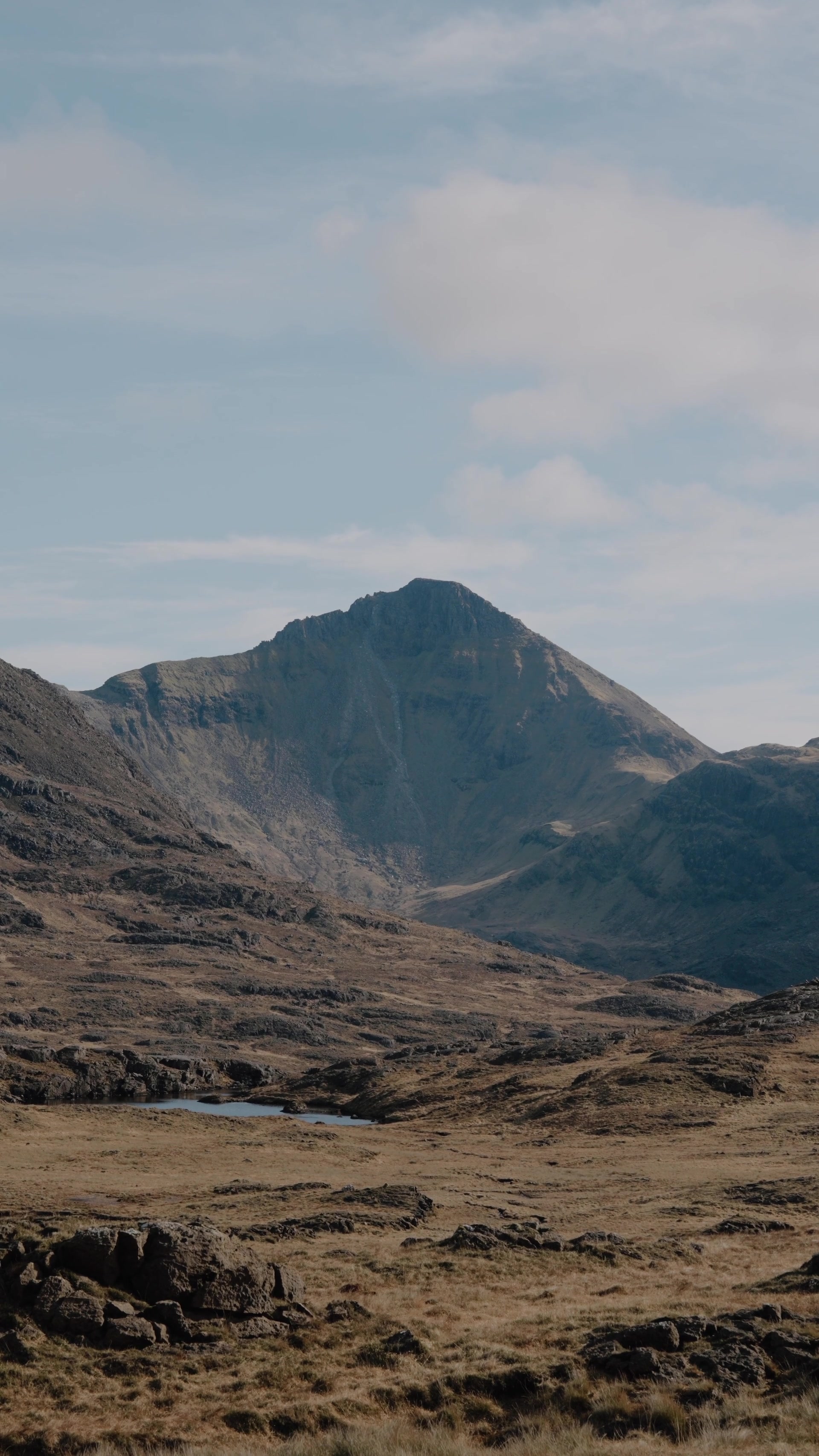 a single sharp mountain peak framed by lower hills, a small loch and rocky moorland below