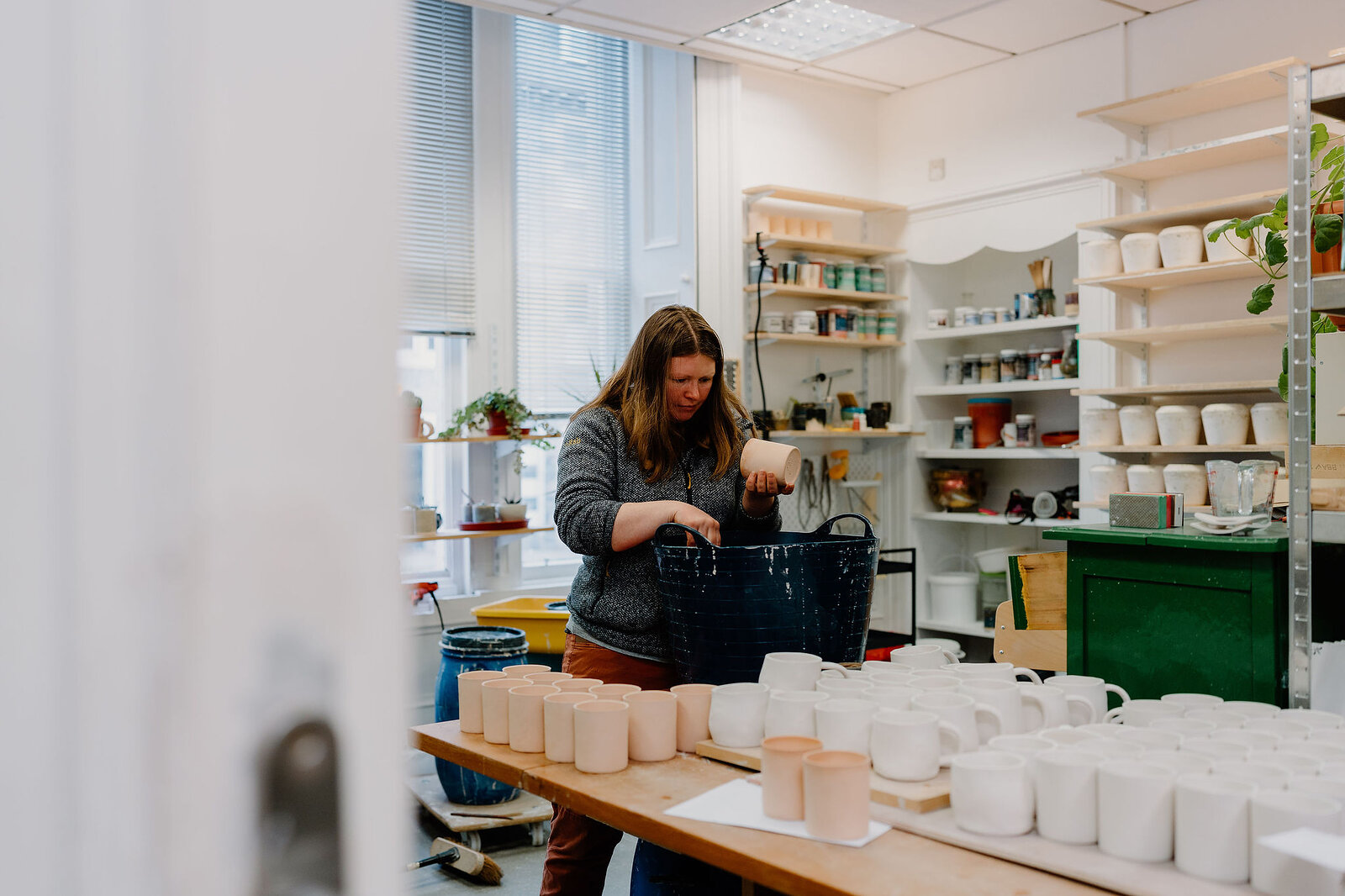 A woman works at a shelf of ceramics beside a green table in a pottery studio, a blue bucket and rows of white cups arranged on the workbench.