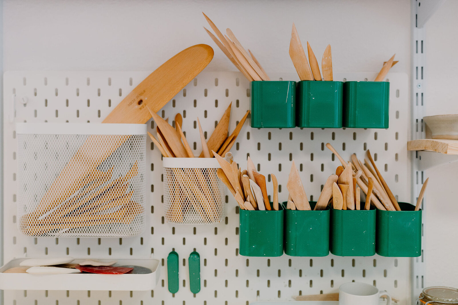 A pegboard wall holds wooden pottery tools and green plastic containers stuffed with brushes and ribs.