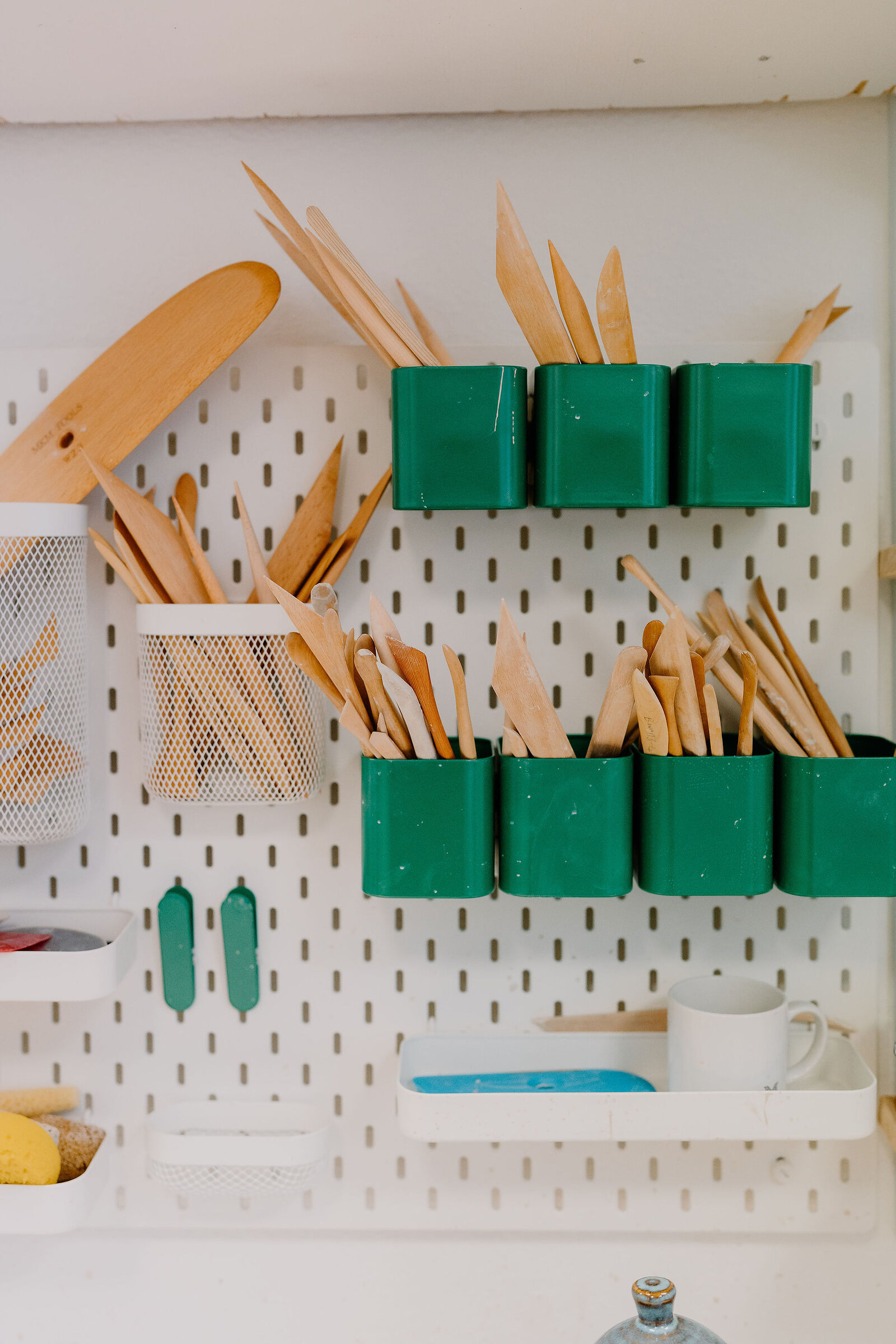 A pegboard wall holds pottery tools in green plastic containers, a mesh basket and a white mug resting on a small tray.