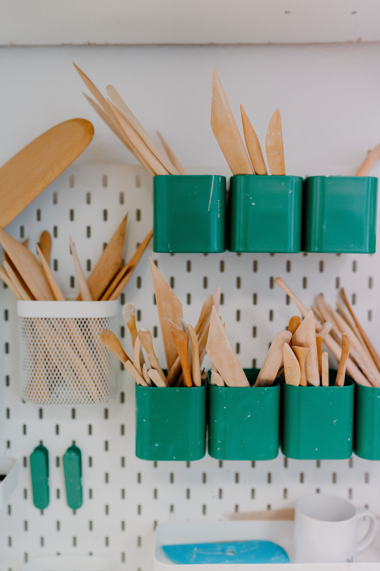 A pegboard wall holds wooden pottery tools in green containers and a mesh basket, wooden spatulas hanging separately to one side.