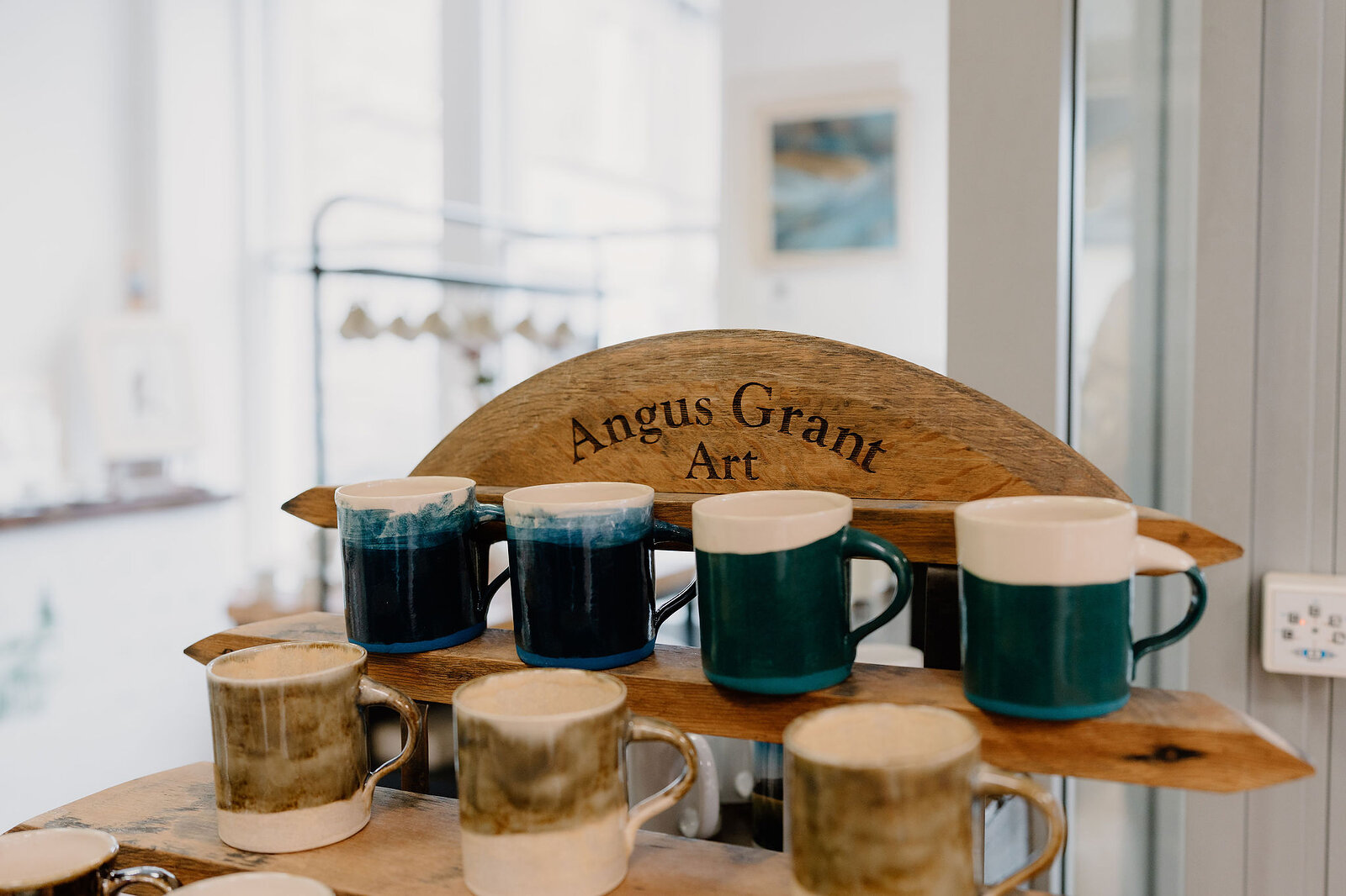 A wooden display rack labelled "Angus Grant Art" holds rows of blue, green and cream ceramic mugs.