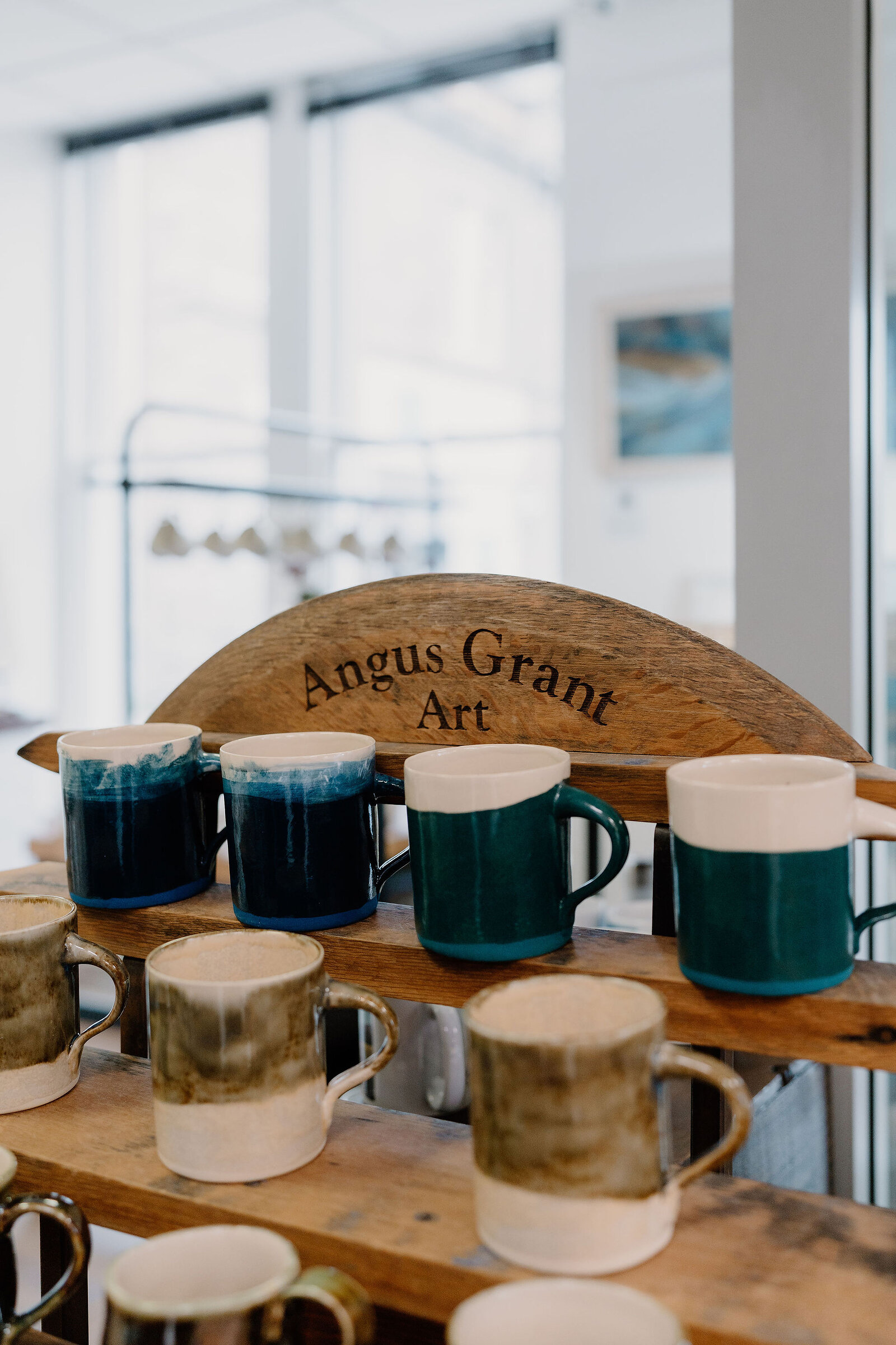 A wooden display rack labelled "Angus Grant Art" holds ceramic mugs in blue and cream glazes.