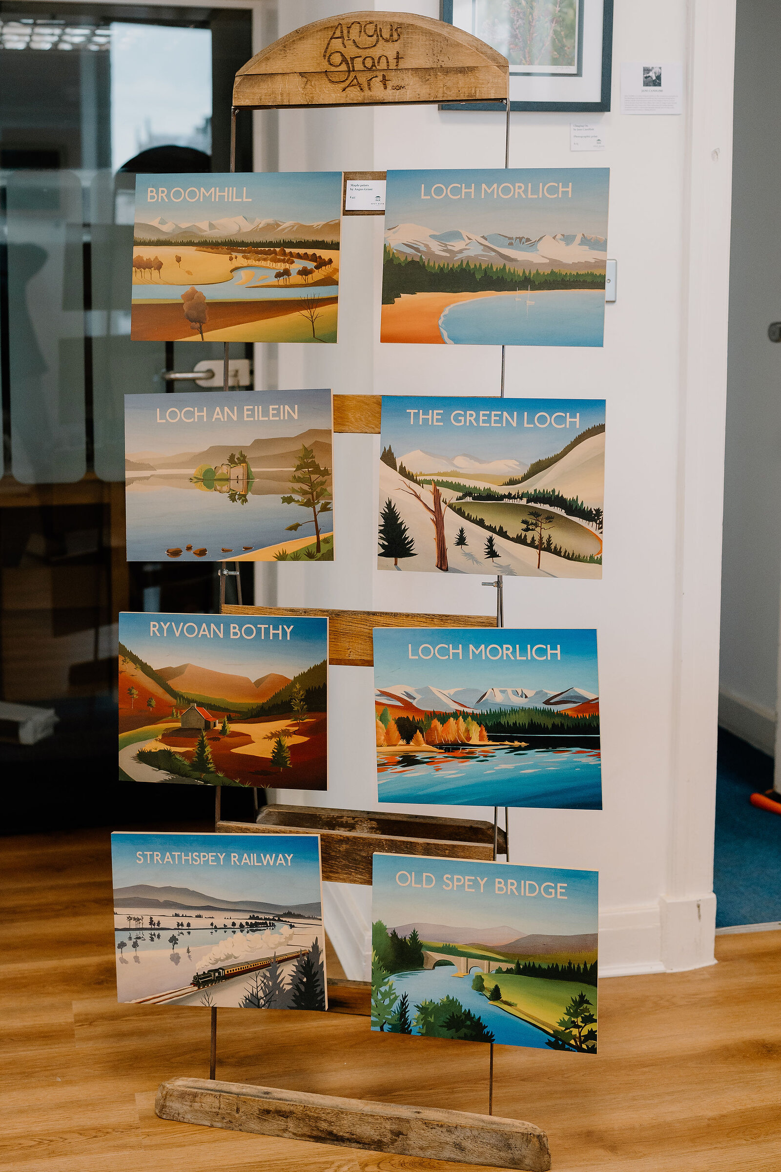 An easel display beneath a sign reading "Angus Grant Art" holds paintings labelled "Broomhill", "Loch Morlich", "Loch An Eilein", "The Green Loch", "Ryvoan Bothy", "Strathspey Railway" and "Old Spey Bridge".