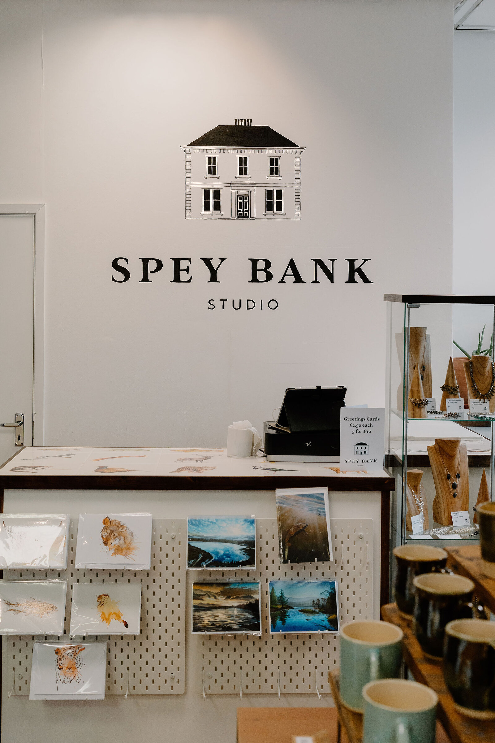 A shop wall carries "SPEY BANK STUDIO" lettering beneath a house logo, a pegboard of prints and greeting cards hanging above a counter of ceramic mugs.