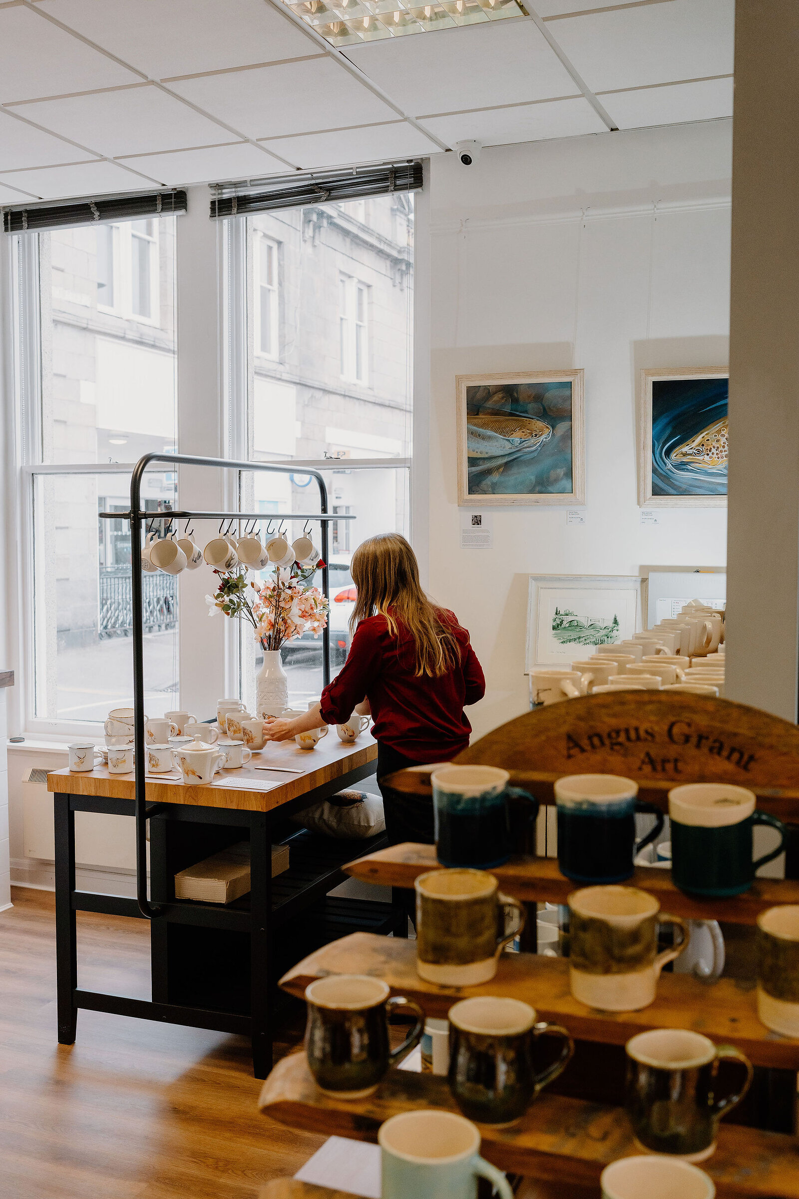 A woman arranges mugs on a table beside a window display in a pottery shop, framed paintings on the wall and a wooden shelf lettered "Angus Grant Art" stacked with glazed mugs in the foreground.