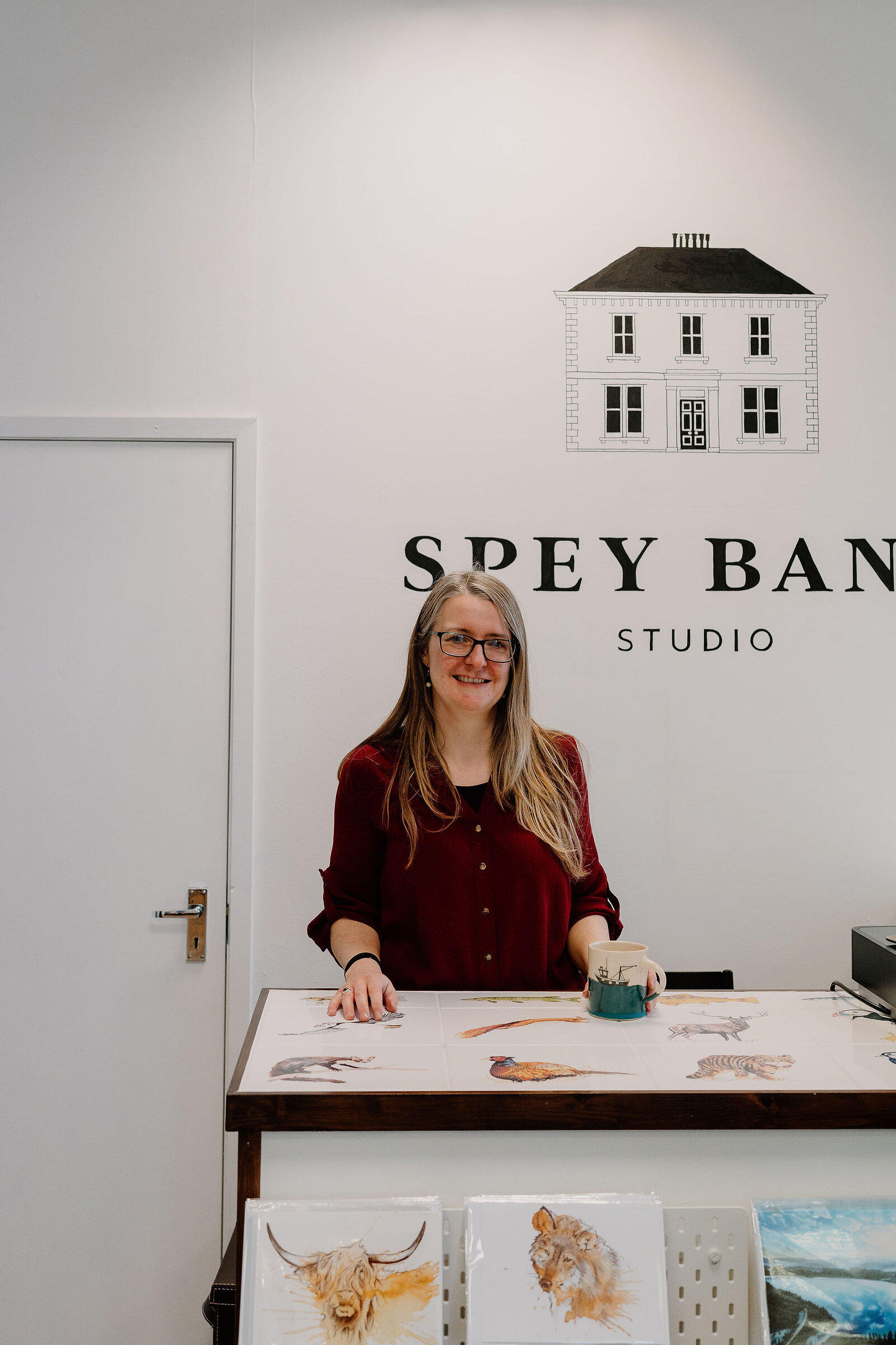 A woman stands smiling behind a counter lettered "Spey Bank Studio", greeting cards printed with wildlife illustrations spread across it and framed prints stacked below.