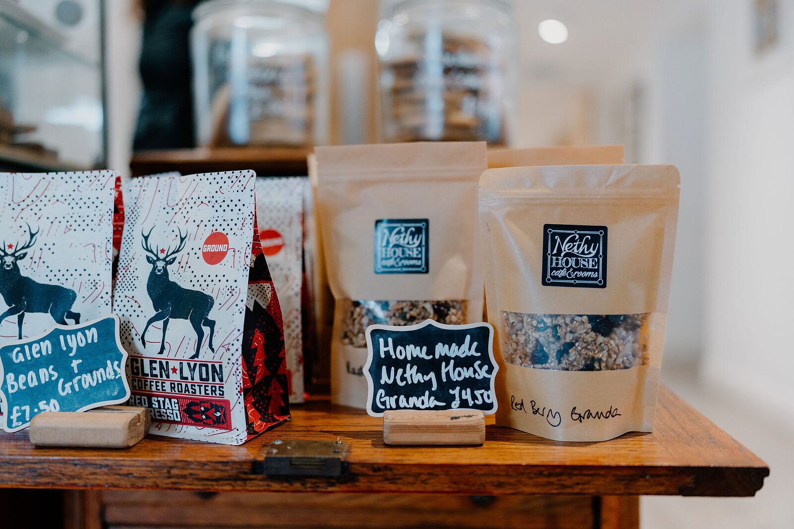 Bags of "Glen Lyon Beans" coffee printed with a stag stand beside "Nethy House Granola" packets and a chalkboard price sign on a wooden counter.