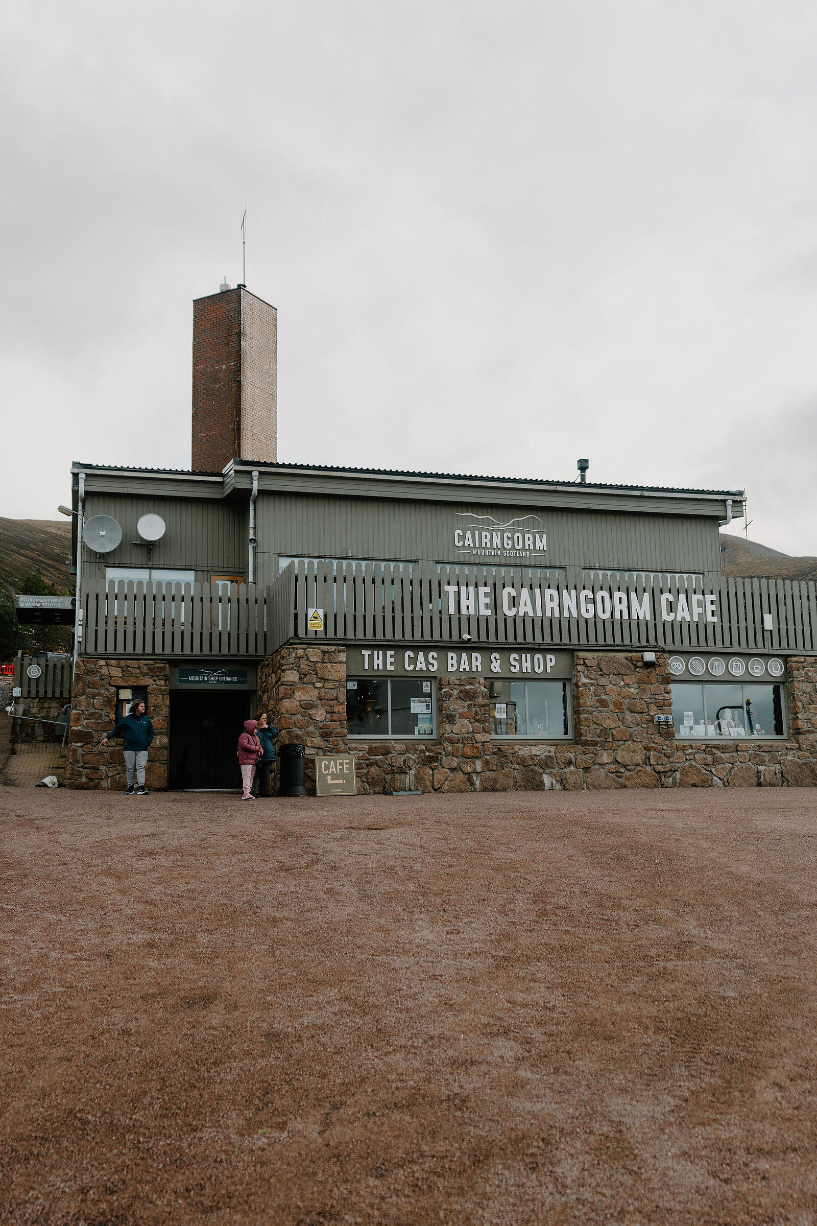 A stone and timber building signed "THE CAIRNGORM CAFE — THE CAS BAR & SHOP" stands on a gravel forecourt, two figures standing near its entrance.