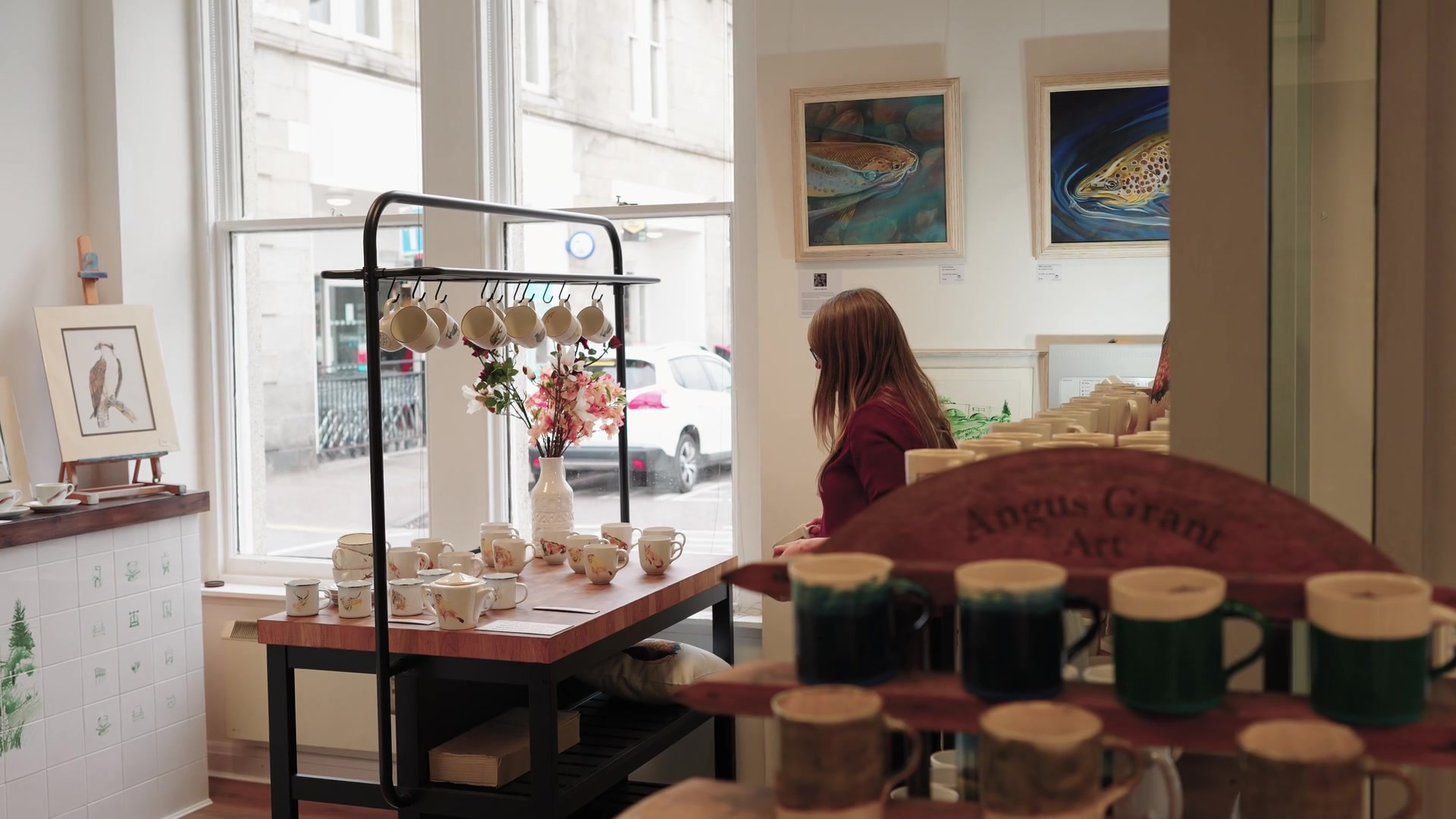A woman browsing framed fish paintings in a gallery hung with mugs and dried flowers