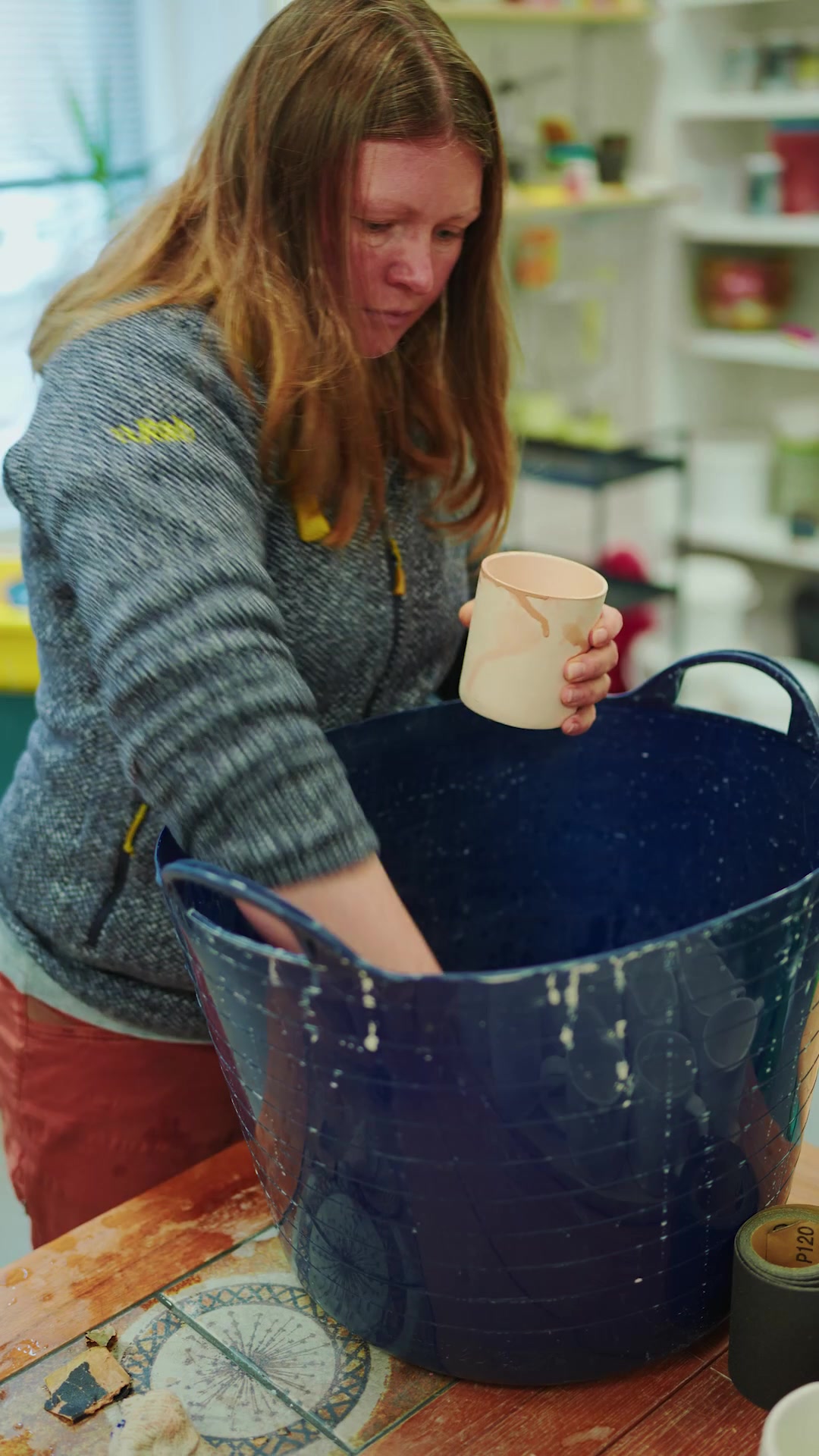 A potter lifting a pale mug from a blue mixing tub in a shop lined with ceramics