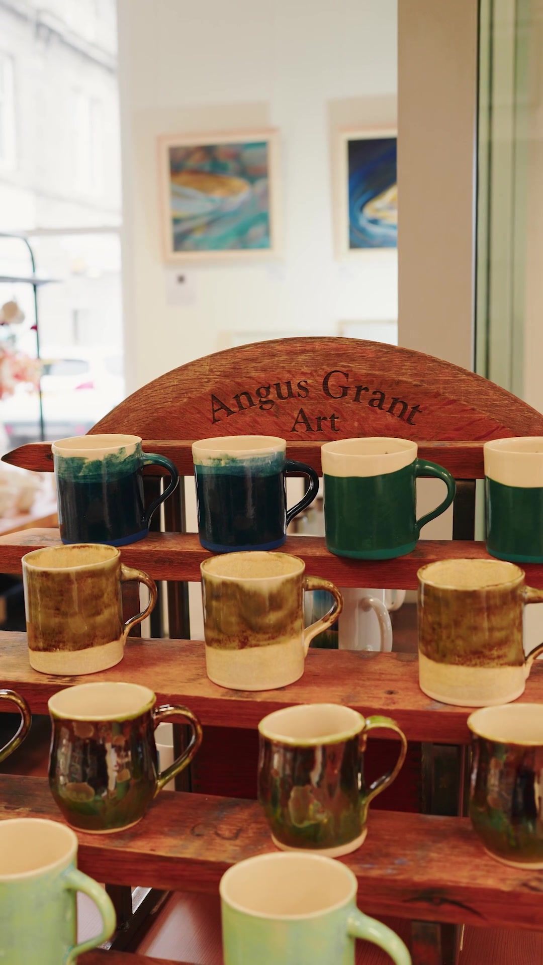 Glazed pottery mugs displayed on a wooden rack beneath an 'Angus Grant Art' sign, framed paintings on the wall behind.