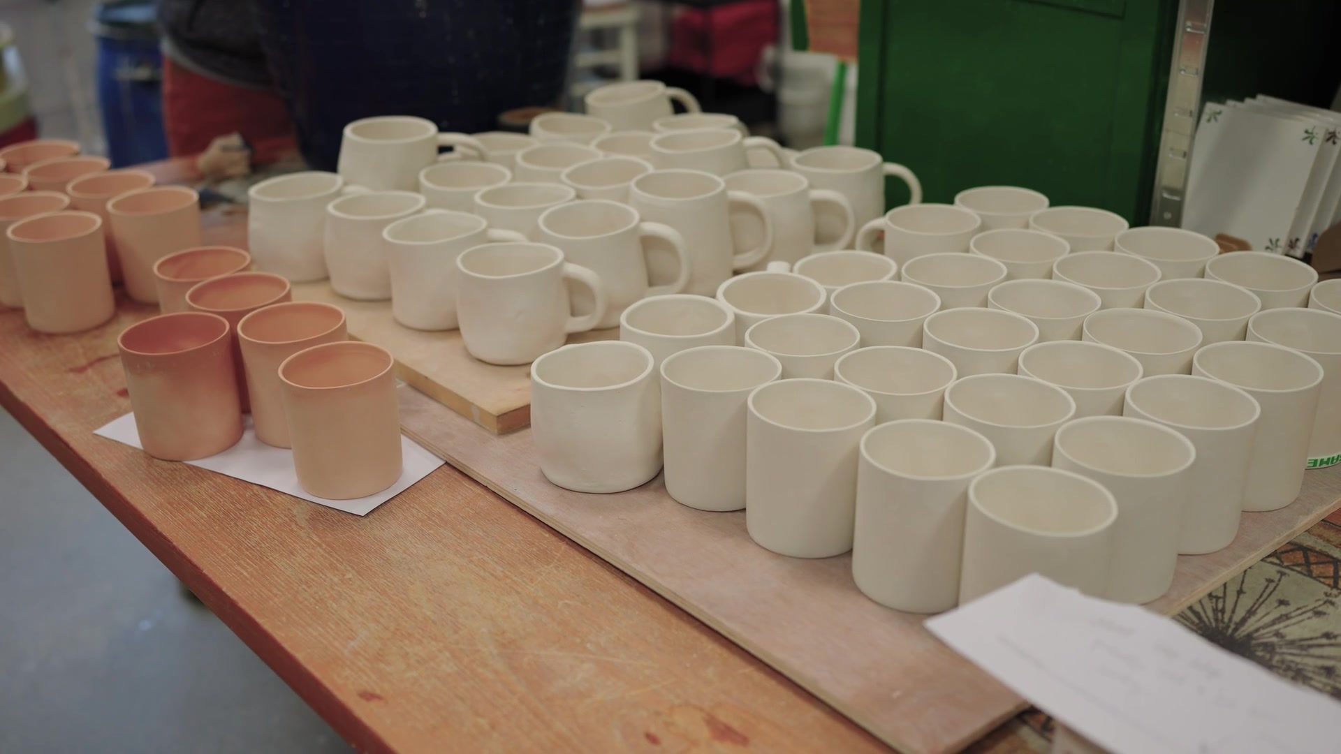 Rows of glazed mugs and cups of varying tones stand ready on a pottery workshop table.