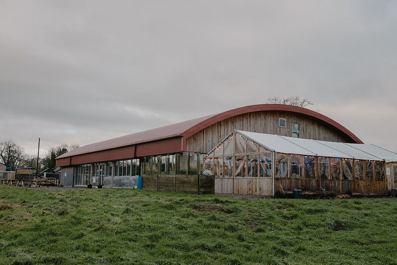 A long barn with a curved red roof and glazed end wall stands in a field, a greenhouse-like extension attached at one side.