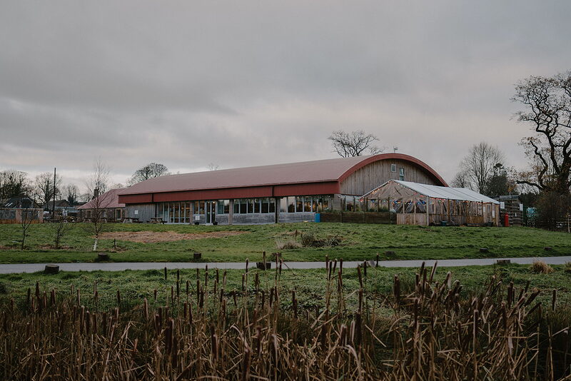 A long barn with a curved red roof stands in a frosted field, reeds growing along a ditch in the foreground.