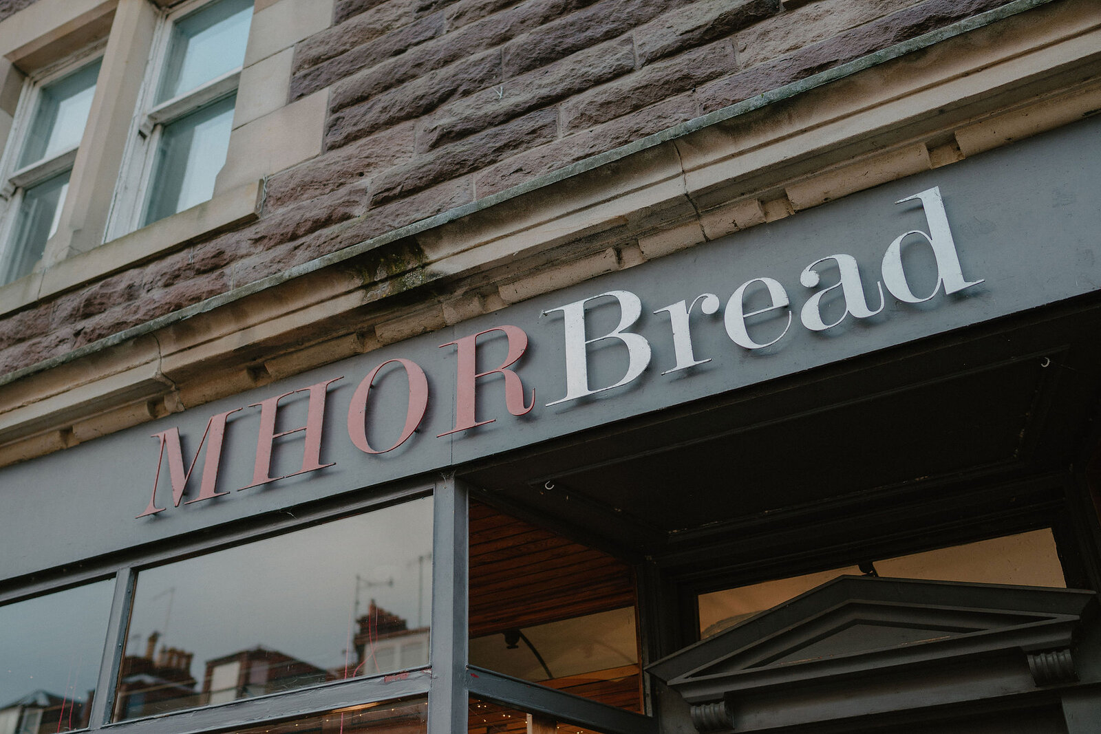 A sign in pink lettering reading 'MHOR Bread' is mounted above a shopfront window on a stone building.
