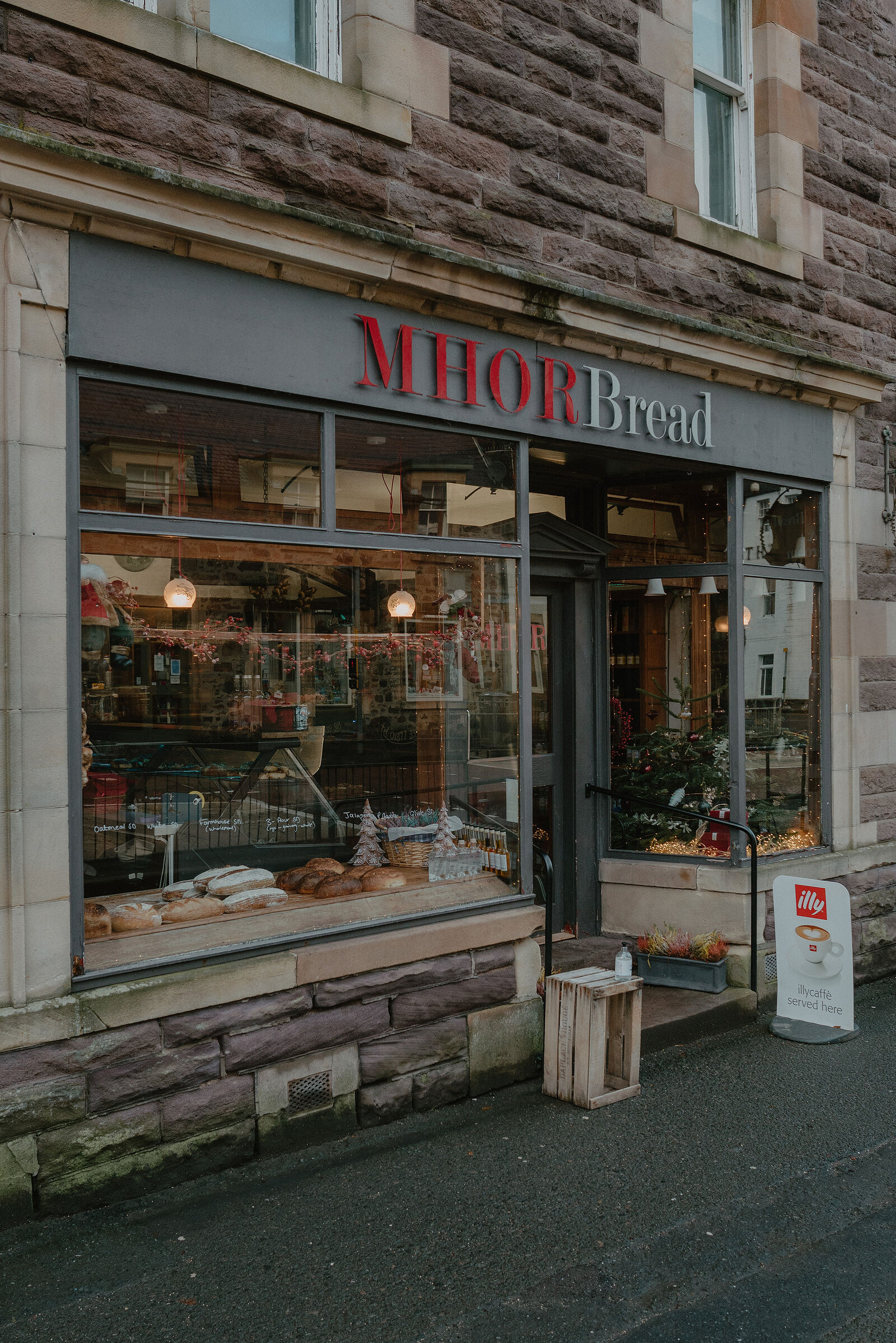 A shopfront lettered 'MHOR Bread' displays loaves in the window beside a small wooden crate and an illy coffee sign on the pavement.
