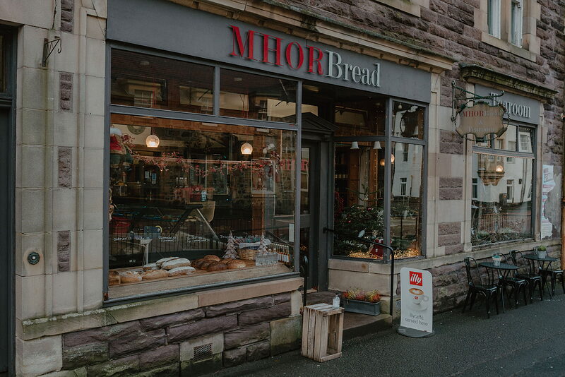 A shopfront lettered 'MHOR Bread' displays loaves and pastries in the window, outdoor tables and chairs set beside the entrance.