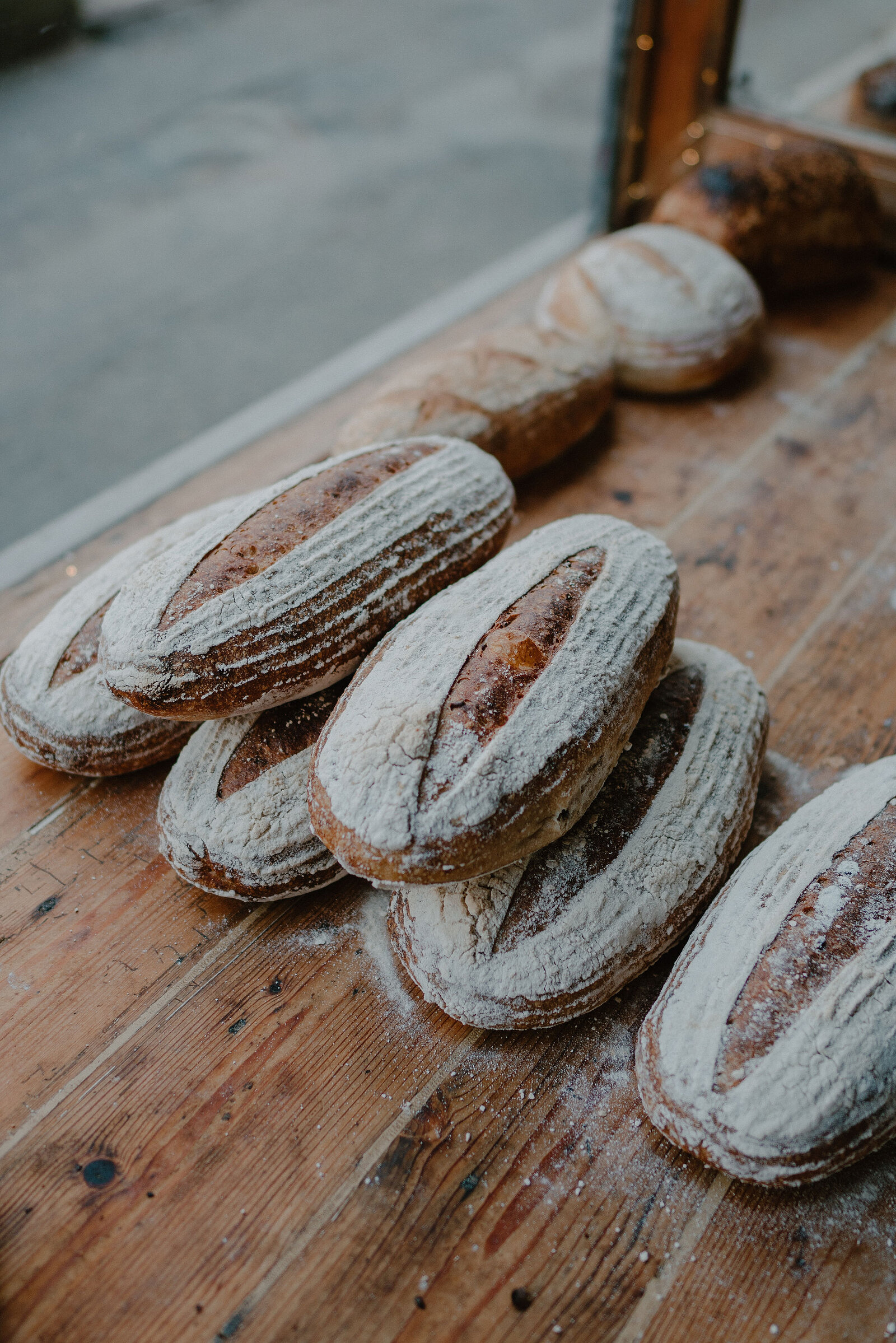 Several oval sourdough loaves dusted with flour sit on a wooden counter beside a shop window.