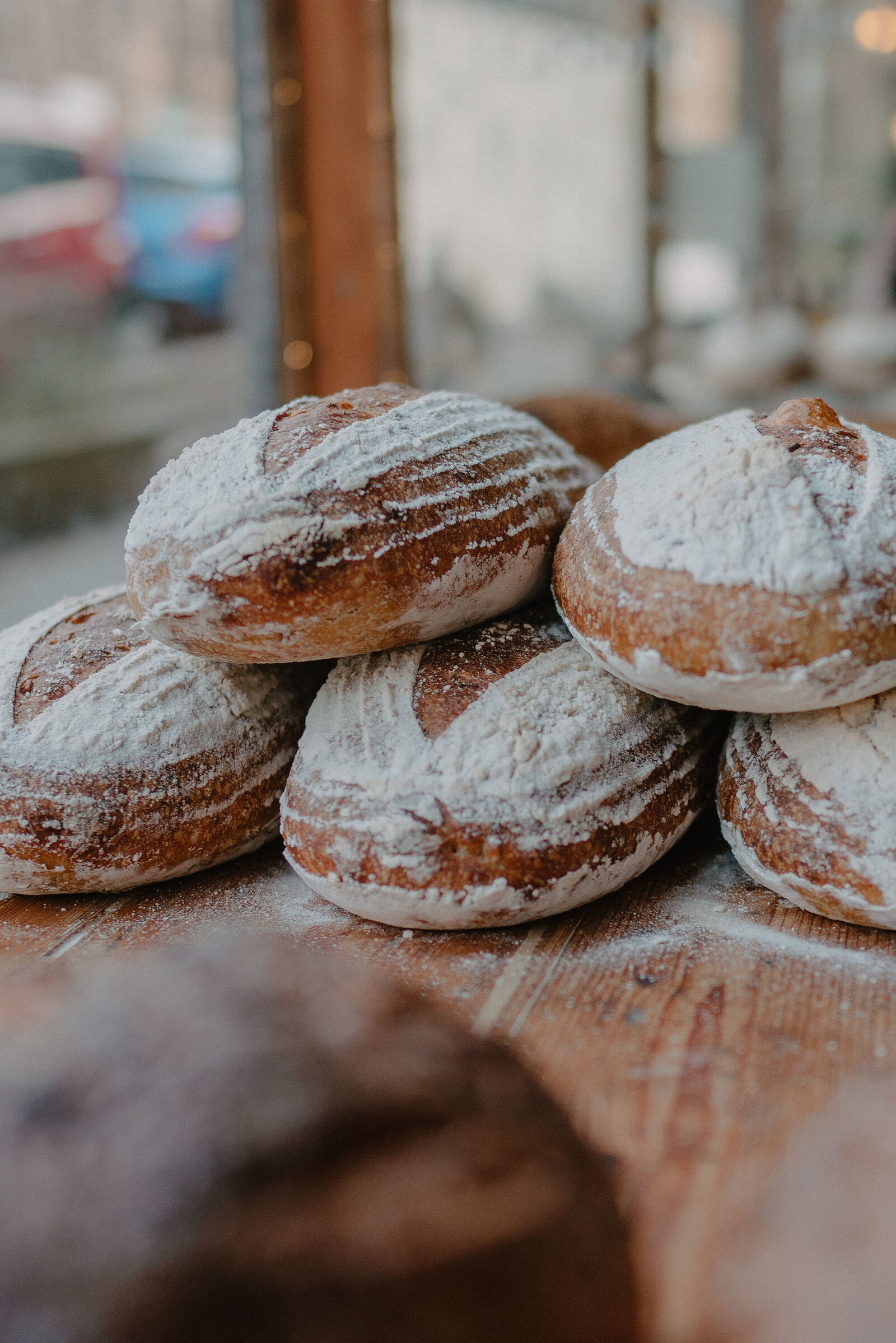 A stack of flour-dusted sourdough loaves sits on a wooden counter beside a bakery window, a car reflected in the glass.