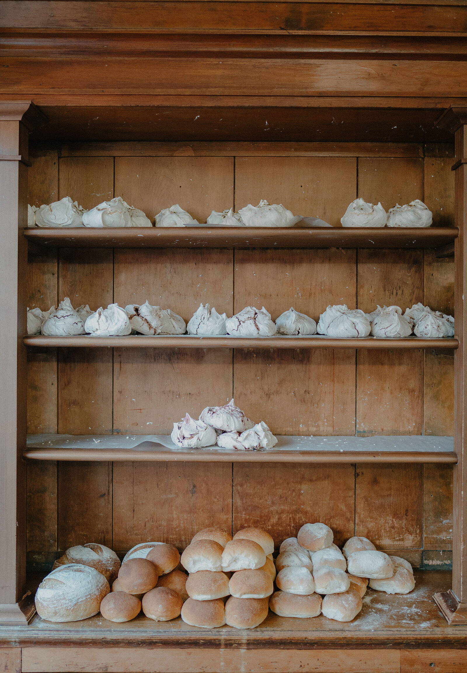 Meringues and bread rolls are arranged on the wooden shelves of an open dresser in a bakery.