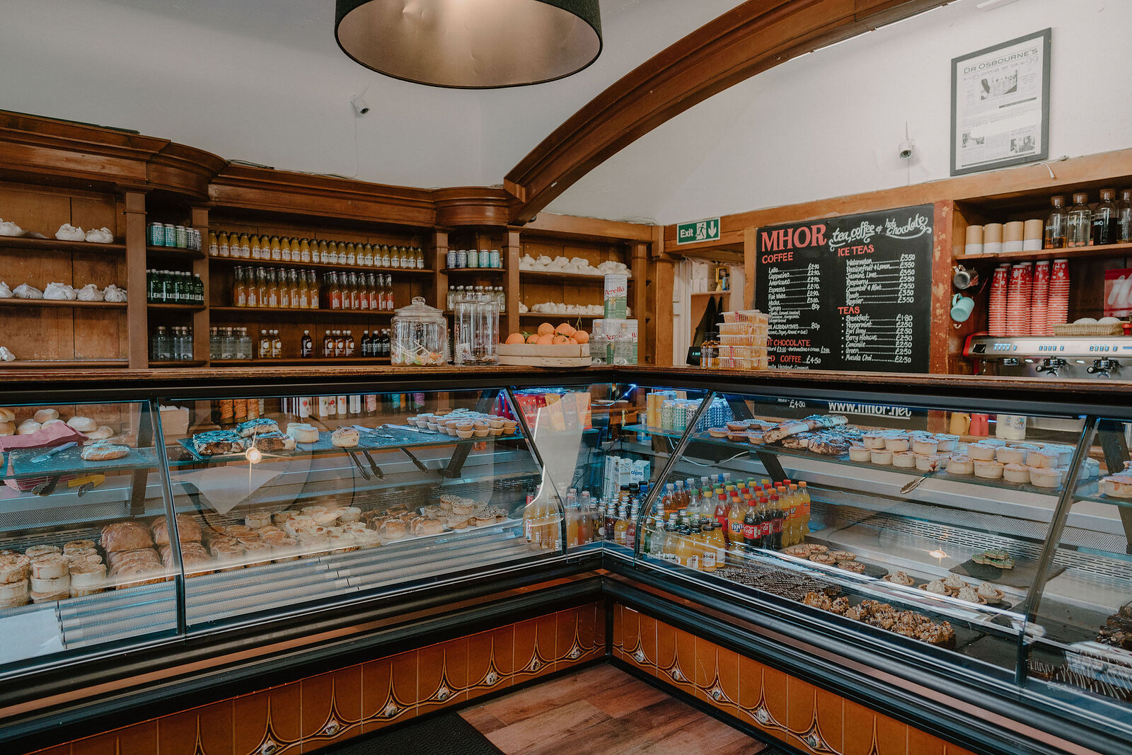 A bakery counter displays cakes, pastries and bread beneath a chalkboard menu reading 'MHOR', shelves of jars and bottles along the wall behind.