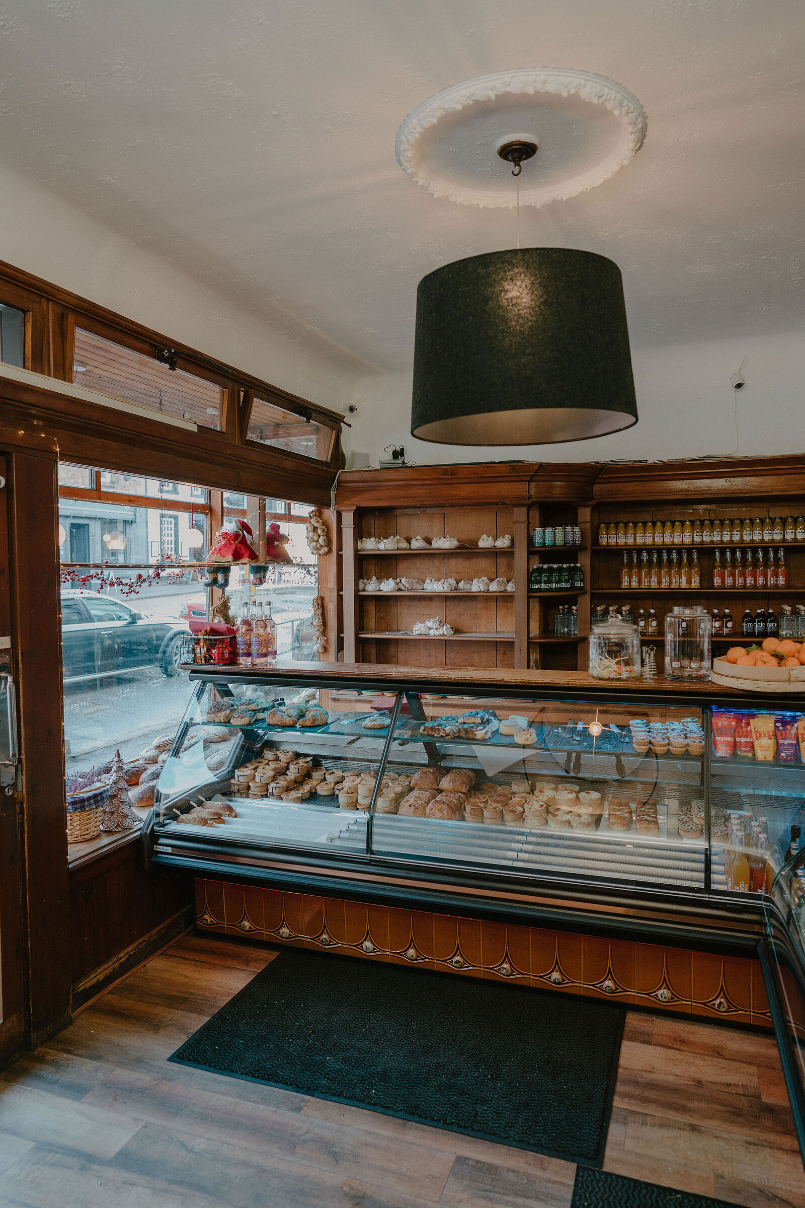 A bakery counter with a curved glass display holds cakes, pastries and bread beneath a wood-panelled shelf of teapots and jars, a dark pendant lampshade overhead.