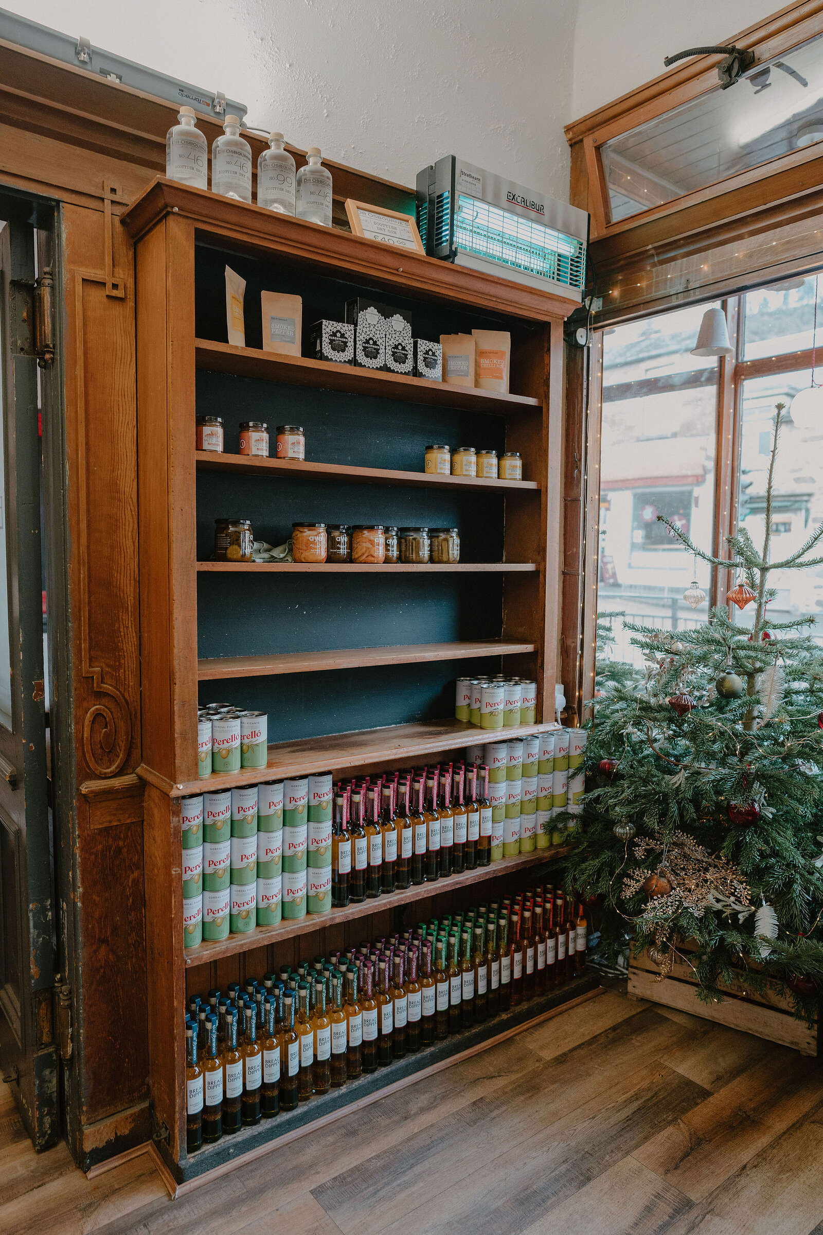 A wooden shelving unit displays candles, jars of preserves and bottled sauces beside a small Christmas tree in a shop window.