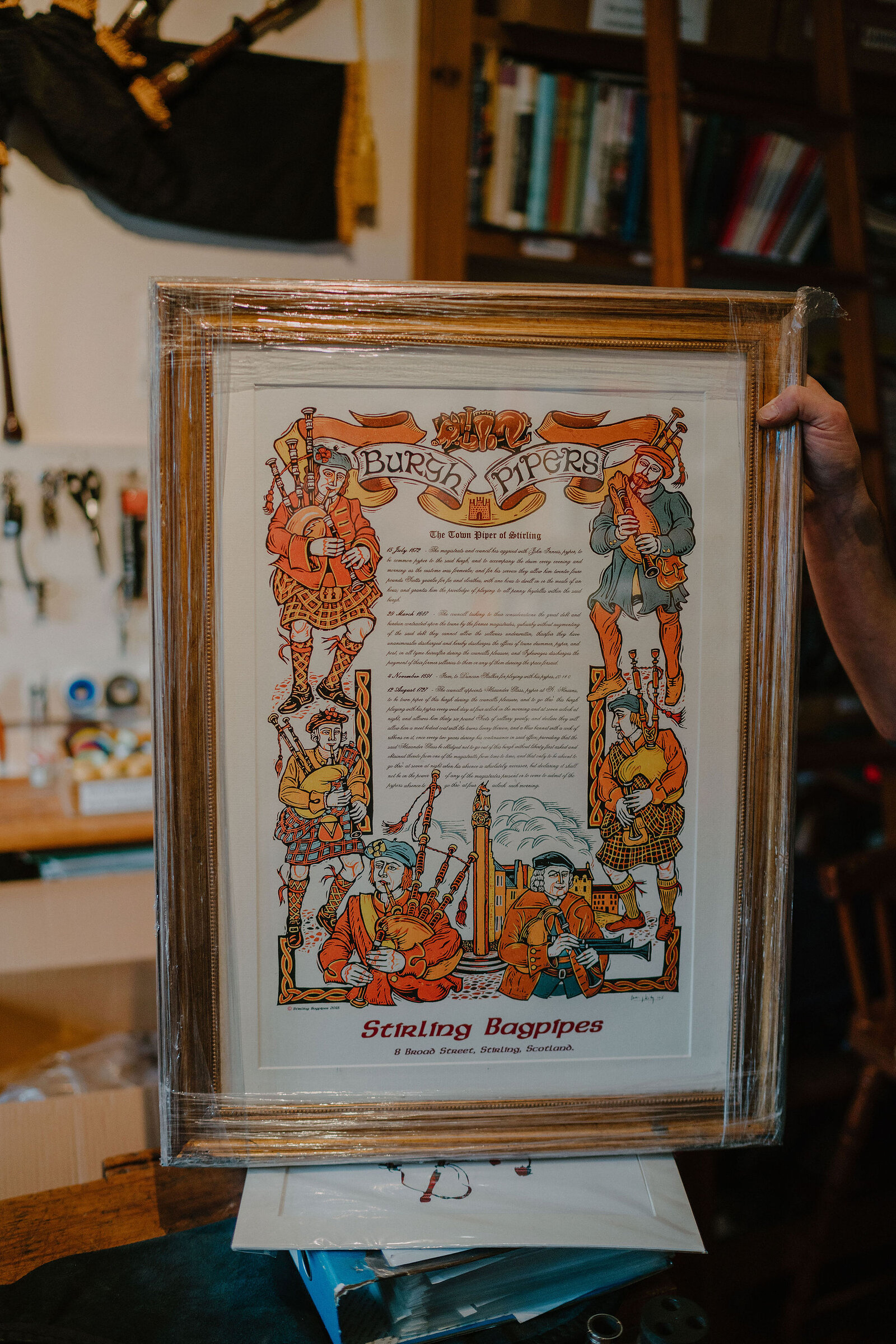 A hand holds up a framed illustrated print titled 'Burgh Pipers, The Toon Pipers of Stirling' branded 'Stirling Bagpipes', shelves of tools and books behind.
