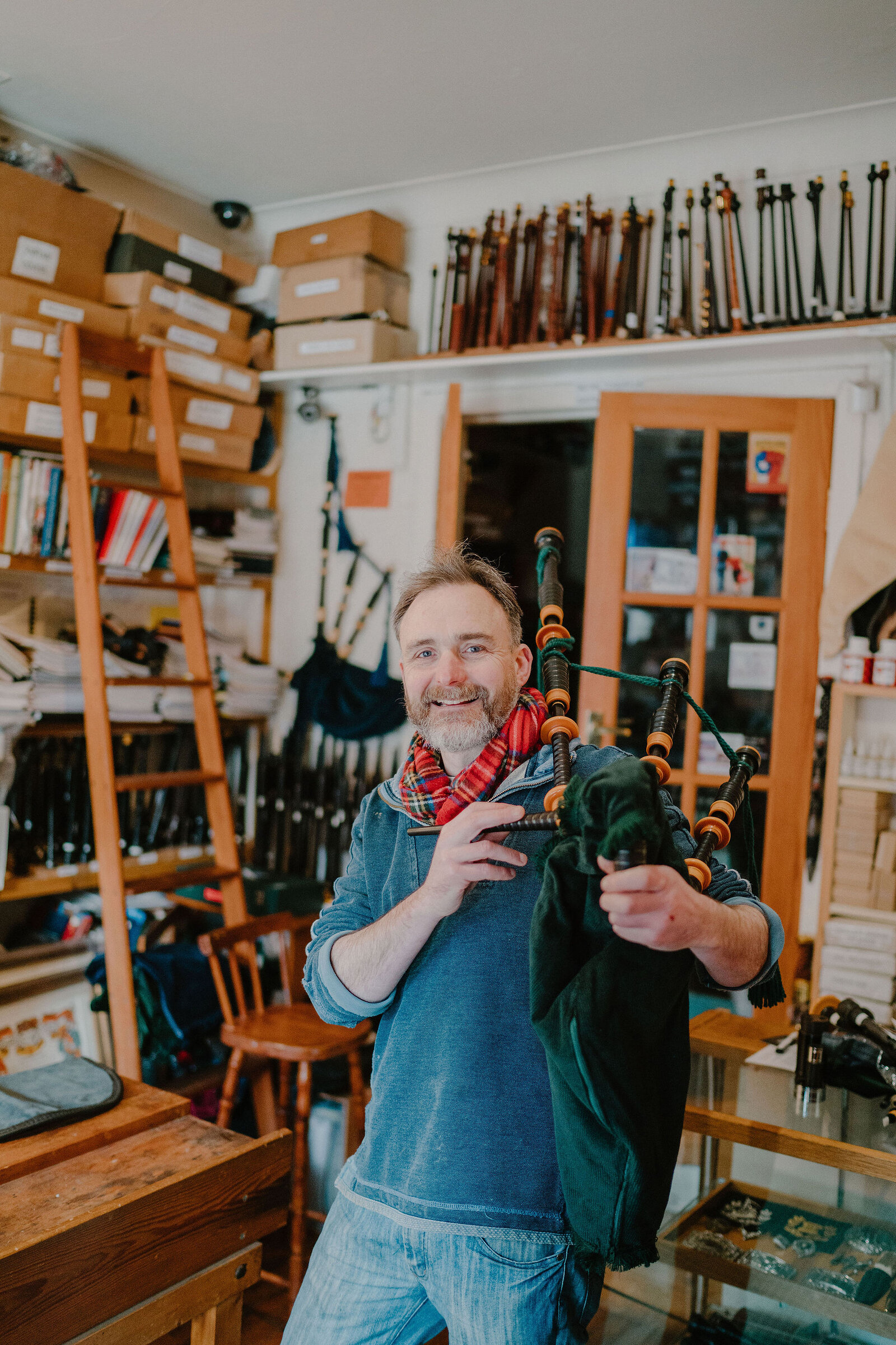 A smiling man in a scarf holds a set of bagpipes, shelves of pipe chanters and boxes of stock filling the workshop behind him.