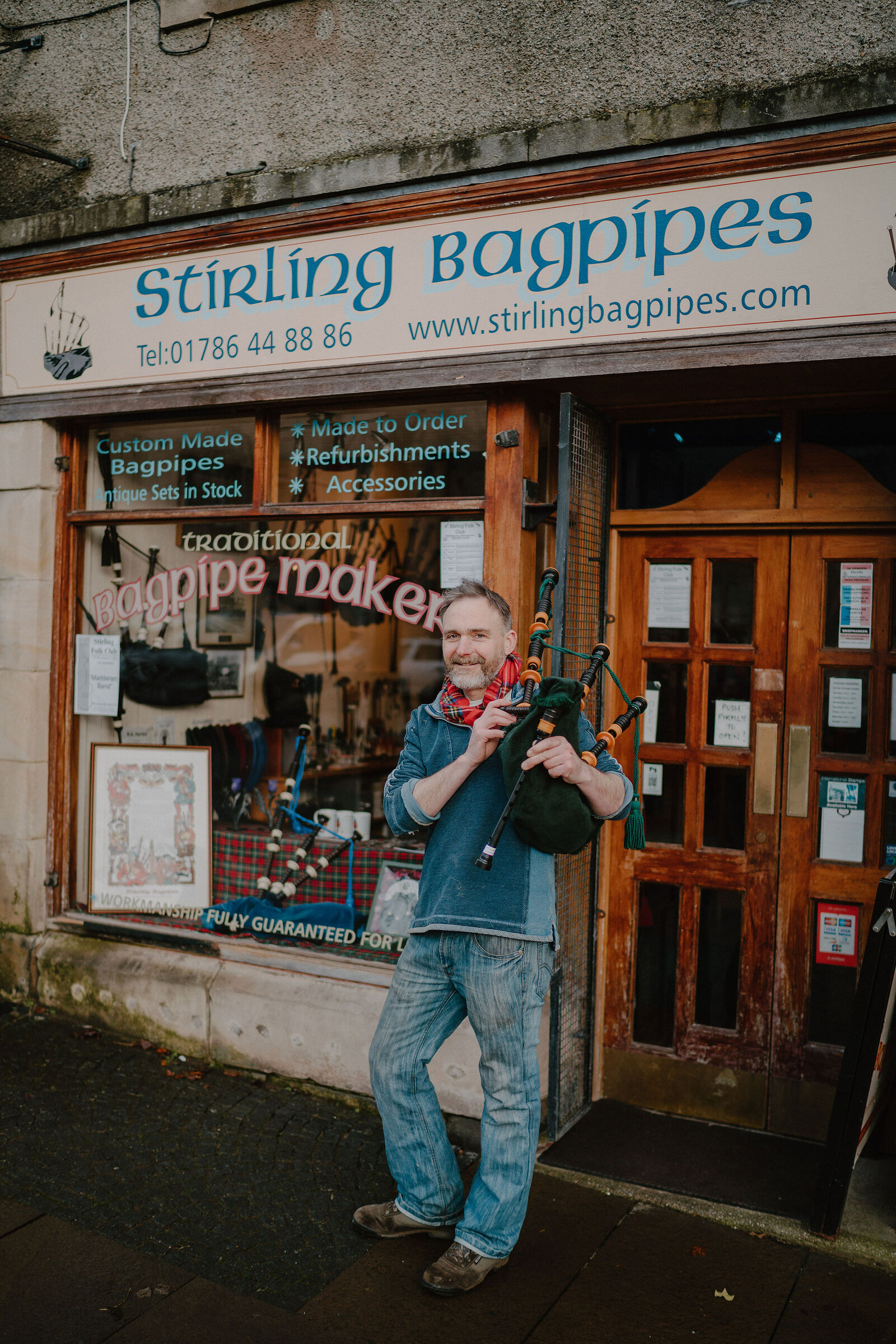 A man plays bagpipes outside a shopfront reading 'Stirling Bagpipes, Custom Made Bagpipes, Made to Order, Refurbishments, Accessories, Traditional Bagpipe Maker'.