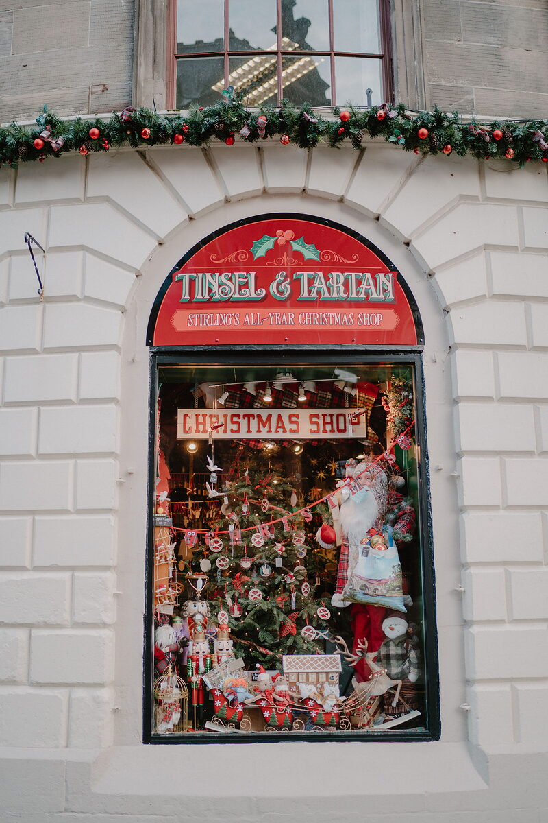 A shop window beneath a sign reading 'Tinsel & Tartan, Stirling's All-Year Christmas Shop' displays a decorated tree, Santa figure and toy sleigh.