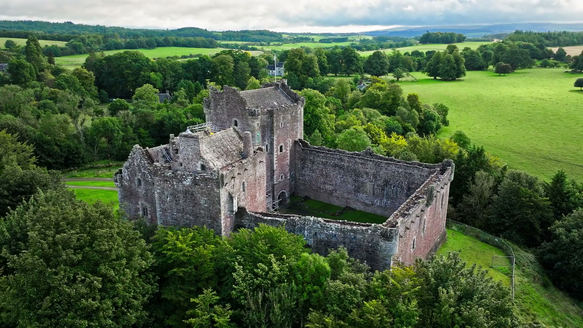 Doune Castle from the air, the gatehouse tower and the walled courtyard among trees, green fields rolling away to the hills beyond.