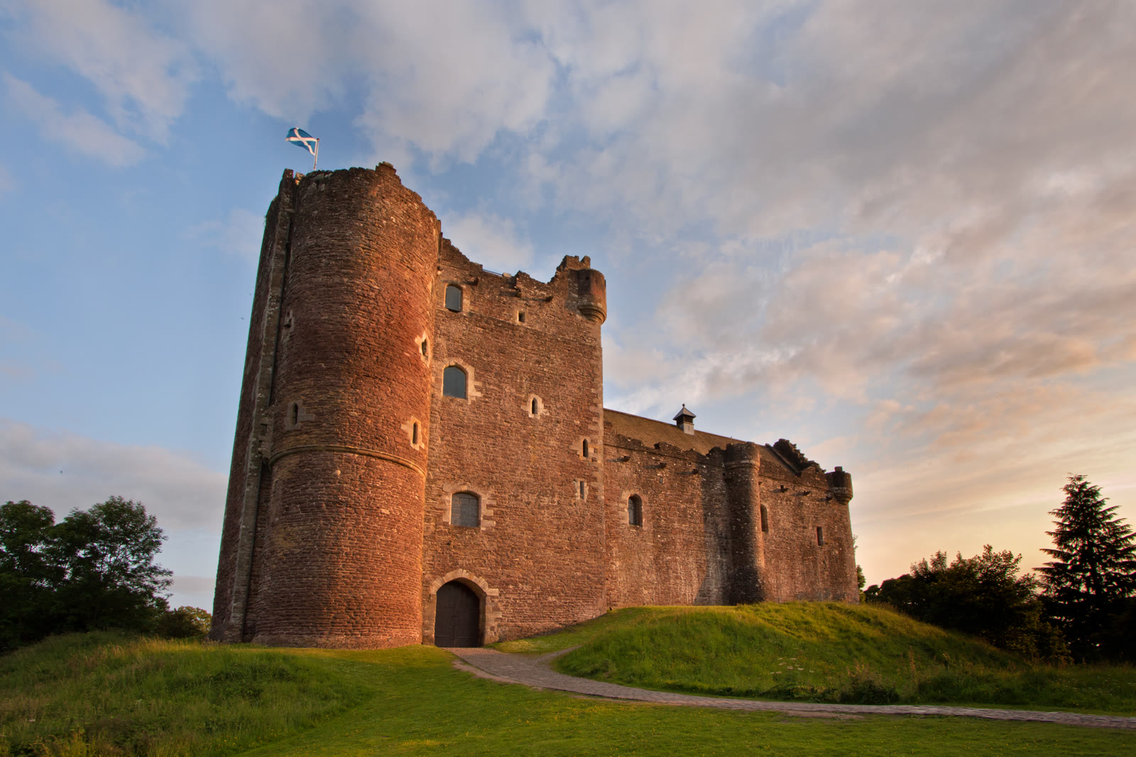 Doune Castle in warm evening light, the round-fronted gatehouse tower and the curtain wall on their green mound, a path curving up to the gate.