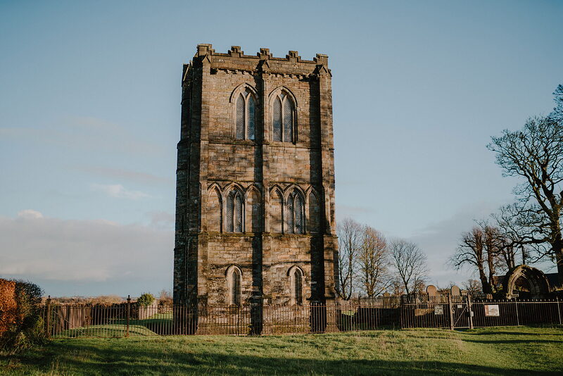 A tall square bell tower with lancet windows and battlements stands alone in a grassy churchyard, bare trees and a fence surrounding it.