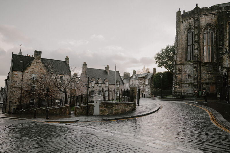 A cobbled street curves past a row of gabled townhouses towards the buttressed corner of a large church, bare trees standing at the junction.