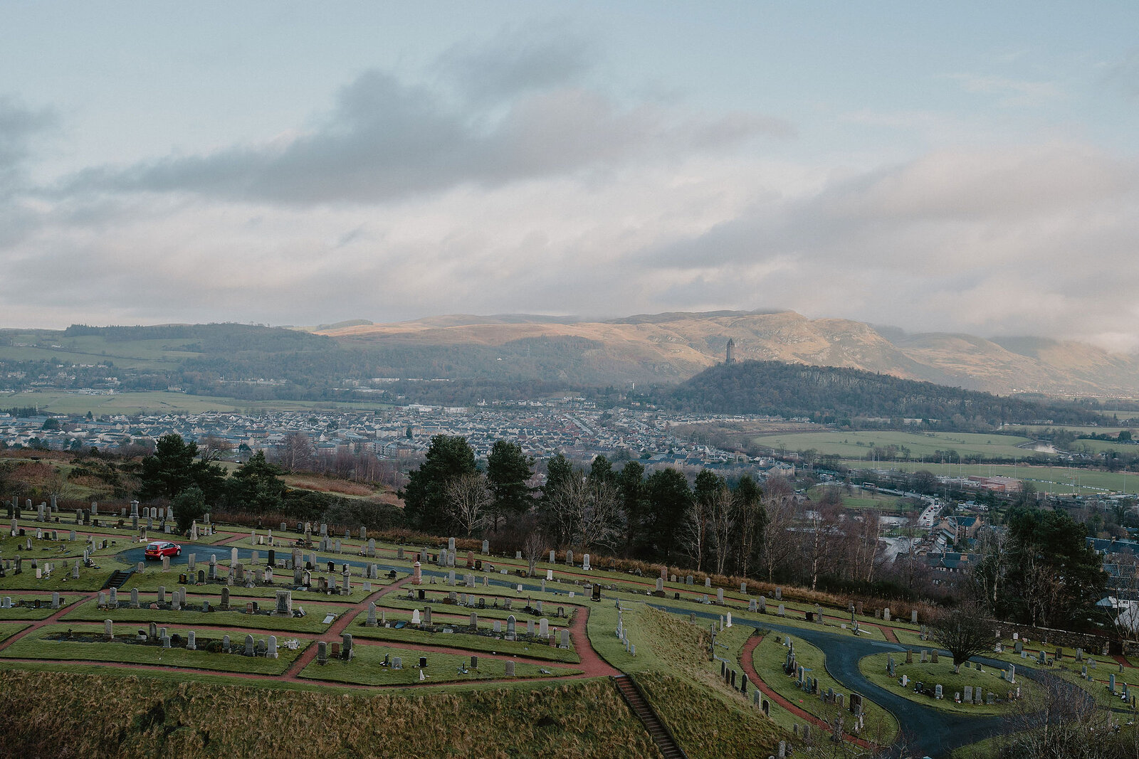 A hillside cemetery with formal garden beds overlooks a town, the Wallace Monument tower visible on a distant wooded hill.