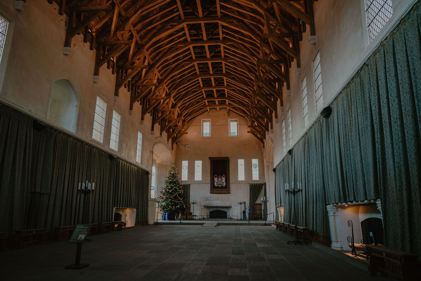 A vast timber-roofed hall has a decorated fireplace and tall windows, a Christmas tree standing at the far end beneath a carved royal crest.