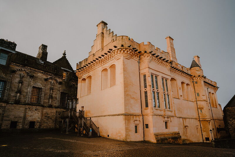 The crow-stepped gables and corner turrets of a royal palace glow in evening light, a stone stair and older tower standing beside it.