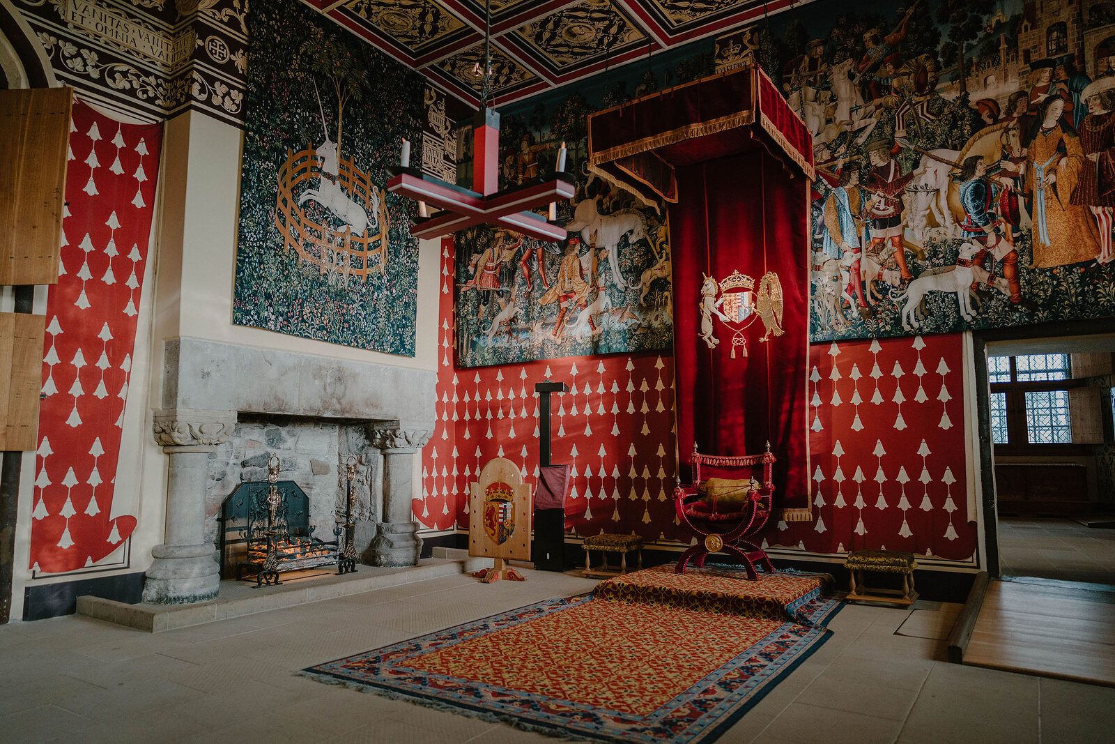 A painted royal apartment with a diamond-patterned ceiling, tapestries and a red canopied throne stands beside a stone fireplace.
