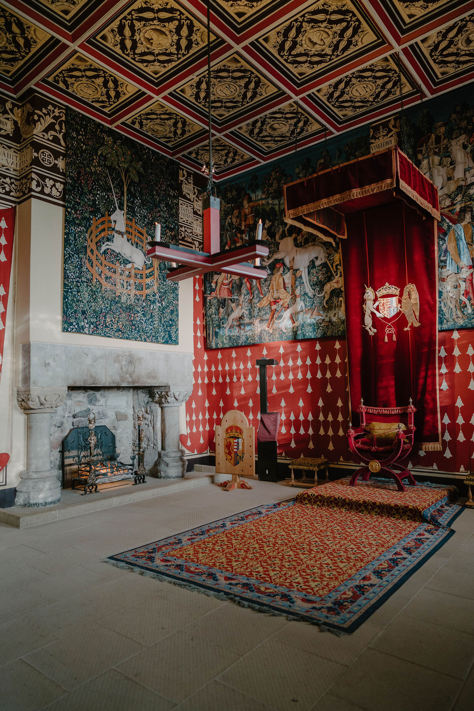 A painted royal apartment with a diamond-patterned ceiling, tapestries and a canopied throne holds a marble fireplace and a patterned carpet.