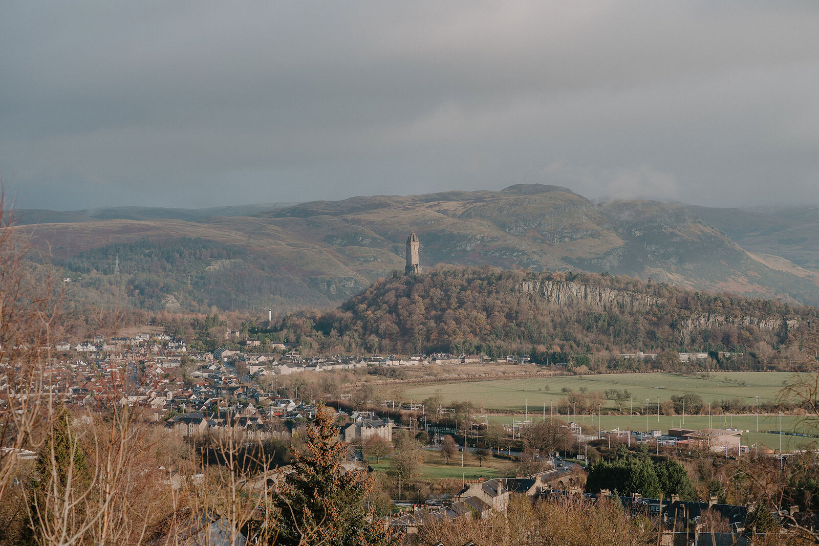 A tall stone monument tower crowns a wooded hill above a town, storm clouds gathering over distant hills.