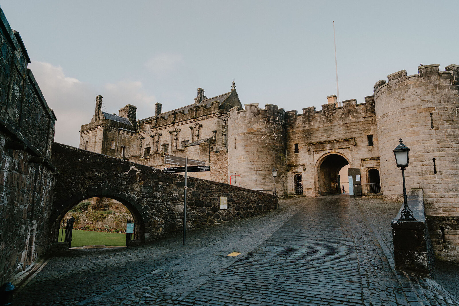 A cobbled forecourt leads through a castle gatehouse flanked by round towers, a stone bridge and archway visible to one side.