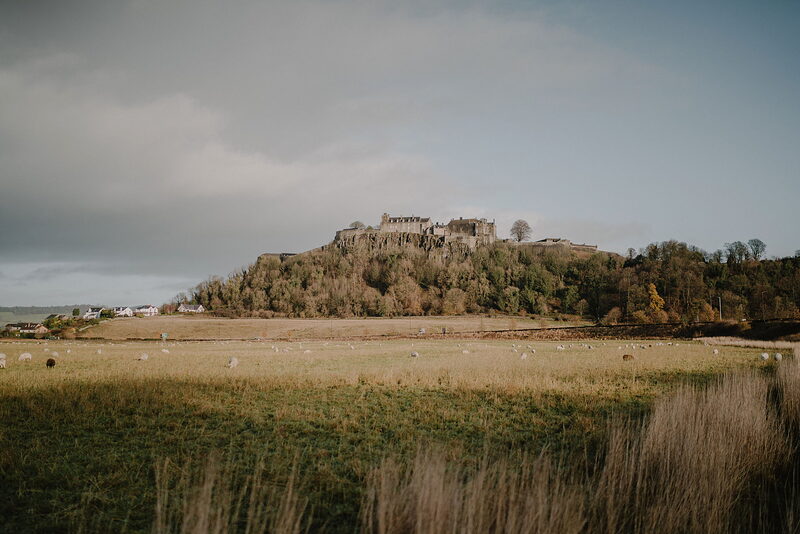 A castle crowns a rocky hill above fields of grazing sheep, hazy hills visible in the distance.