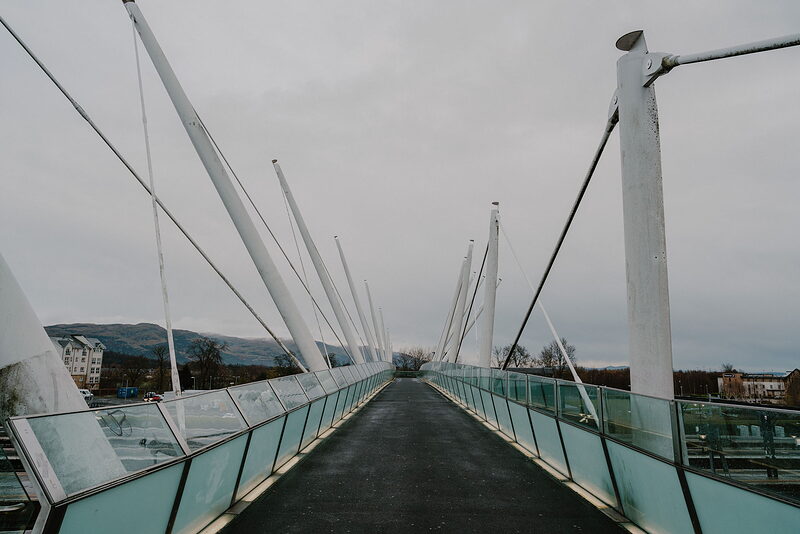 A modern pedestrian bridge with angled mast-like white struts and glass balustrades stretches toward a distant town and hills.
