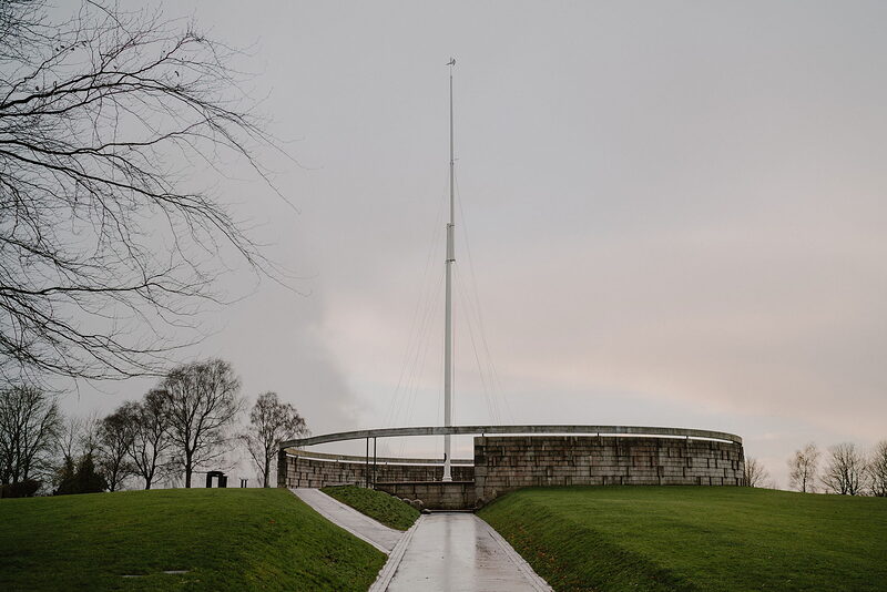 A tall white flagpole rises from a grass mound encircled by a low stone wall, bare trees lining the path beside it.